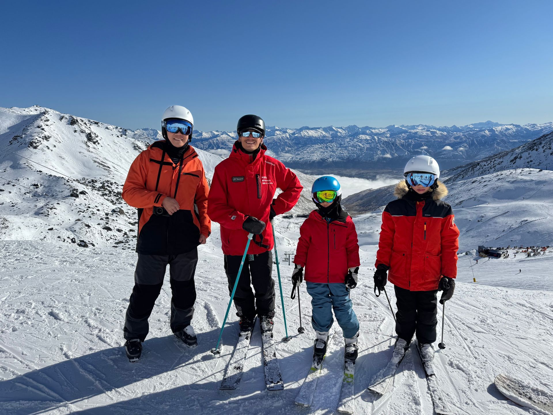 Four people in colorful ski gear and helmets stand together on a snowy mountain summit on a clear day.