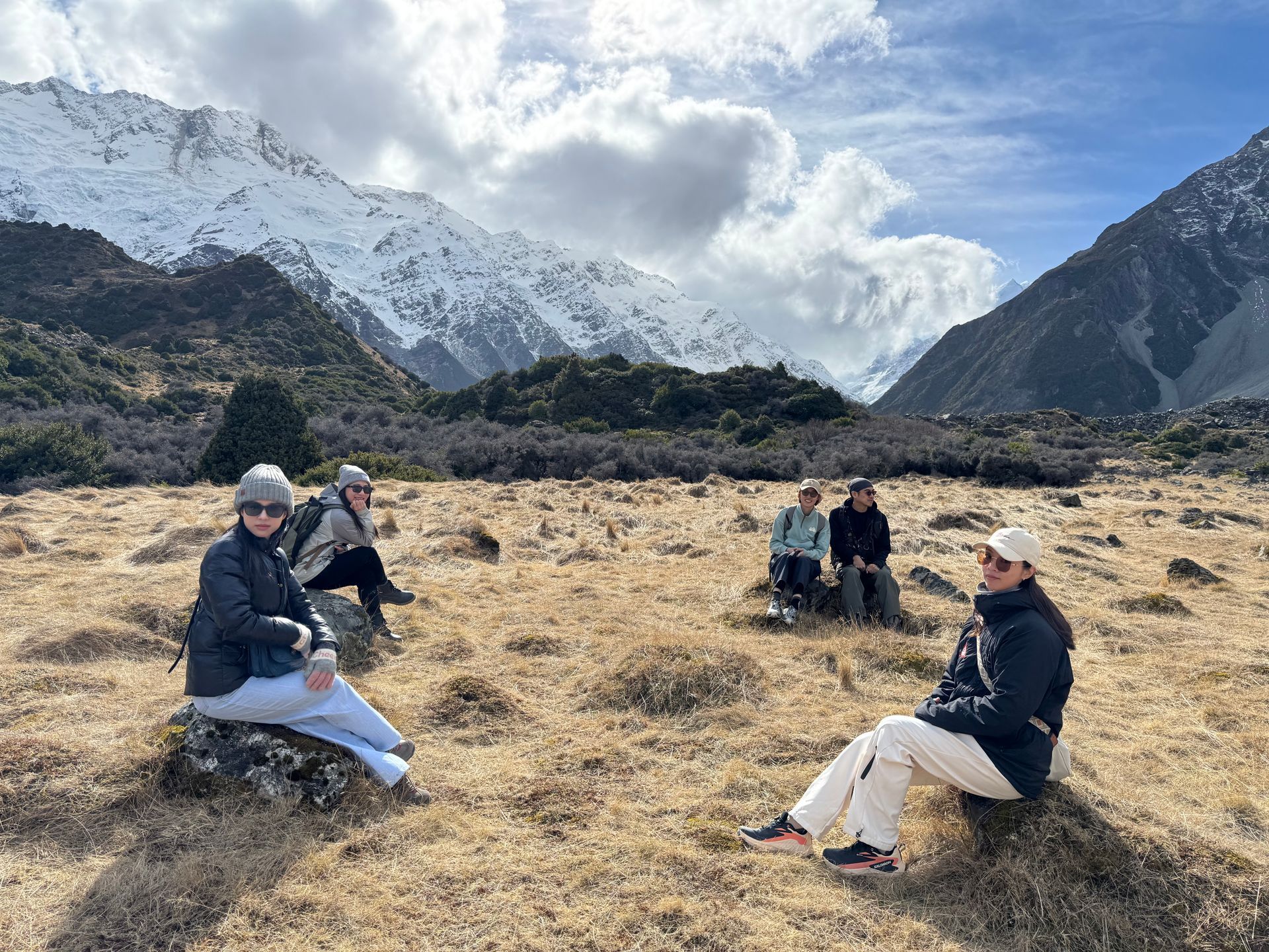 Four people sit on tussock grass in an open field, with snow-covered mountains and a cloudy blue sky in the background.