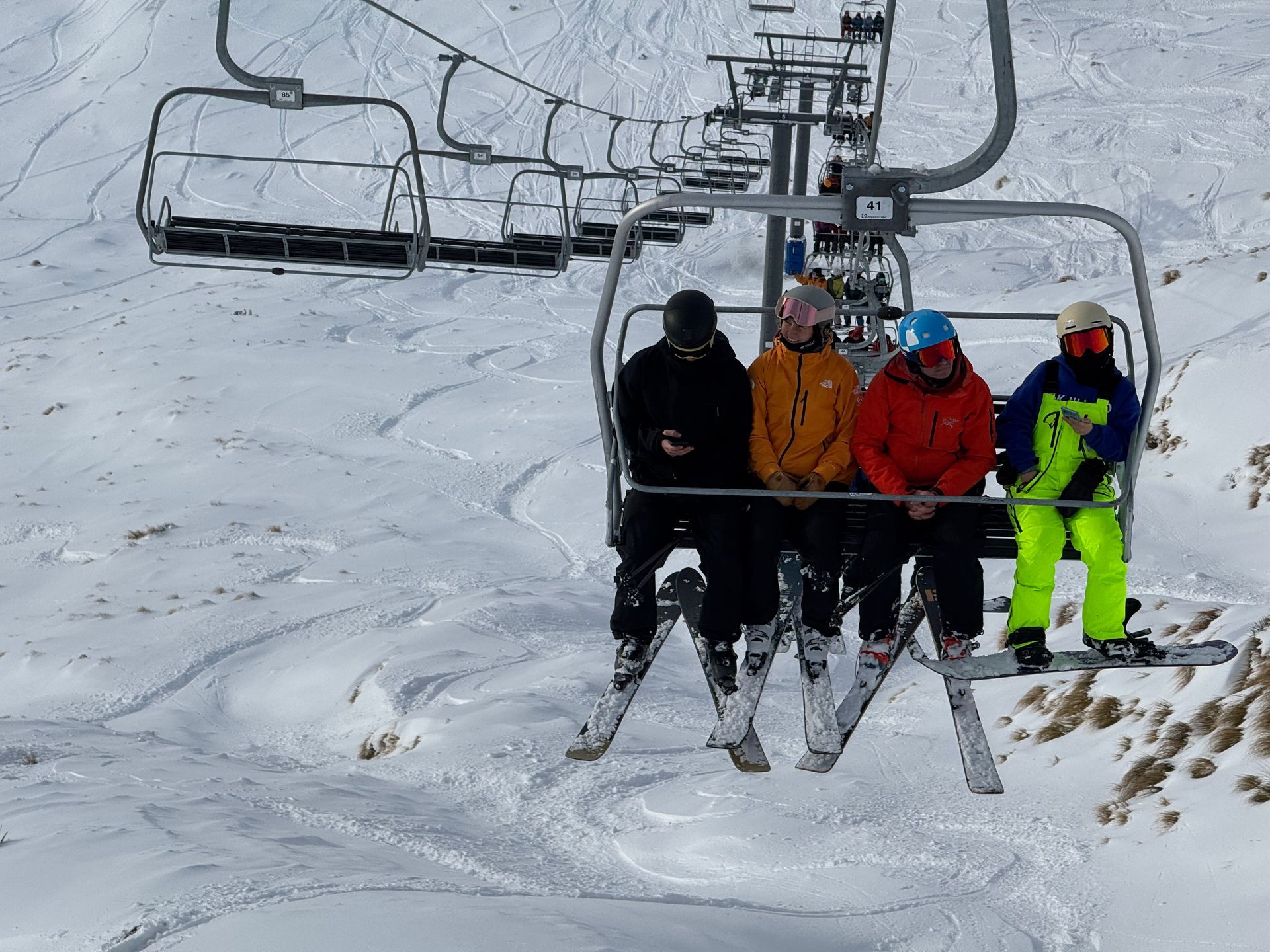 Four skiers ride a chairlift up a snow-covered mountain, wearing bright jackets including yellow, orange, and lime green.