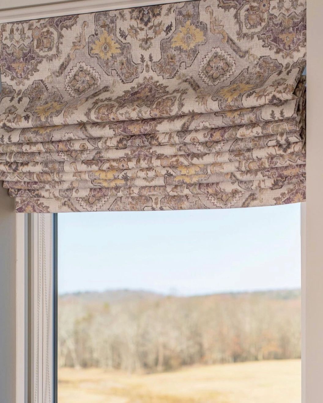 A window with a roman shade on it and a view of a field.