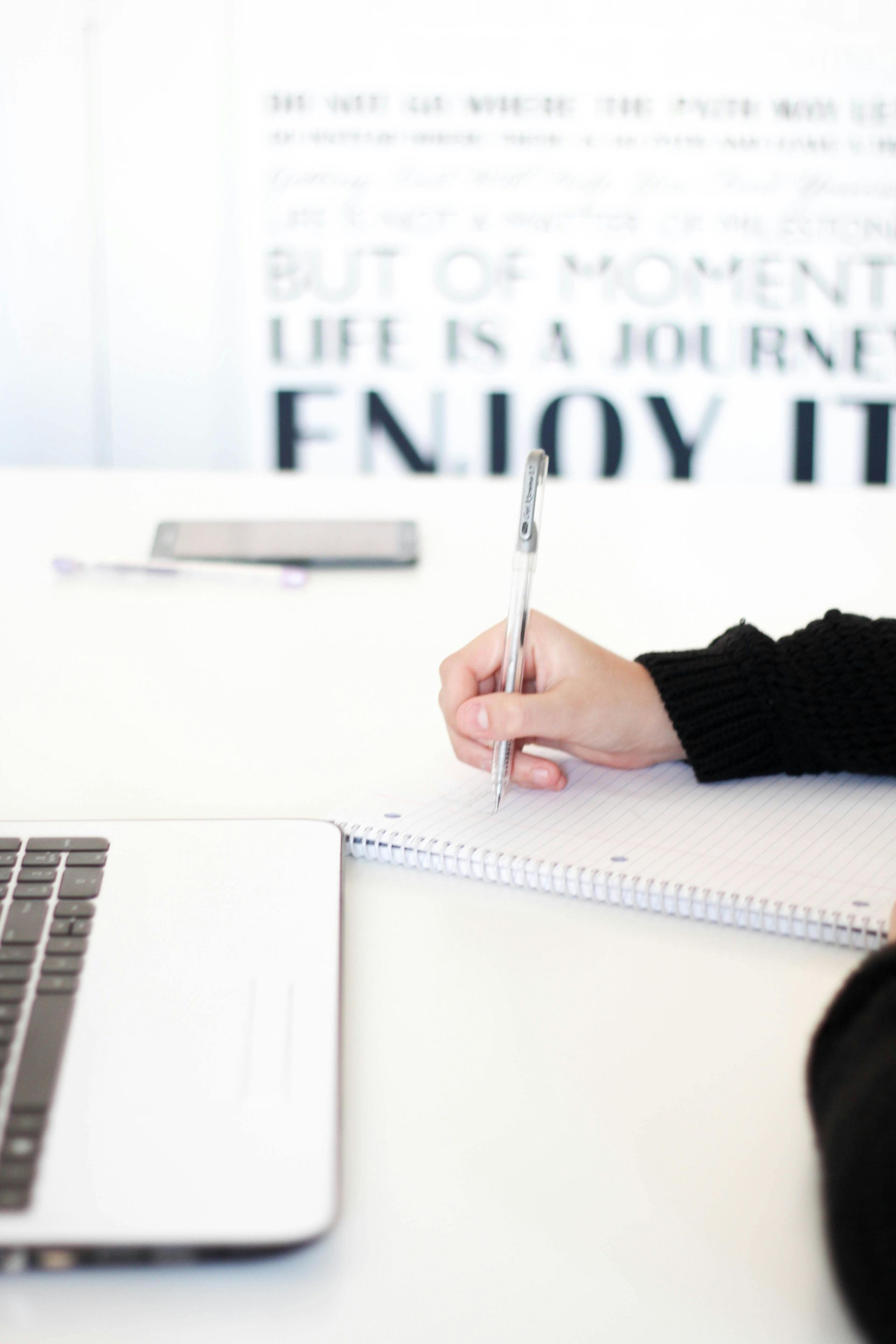 A person is writing in a notebook next to a laptop