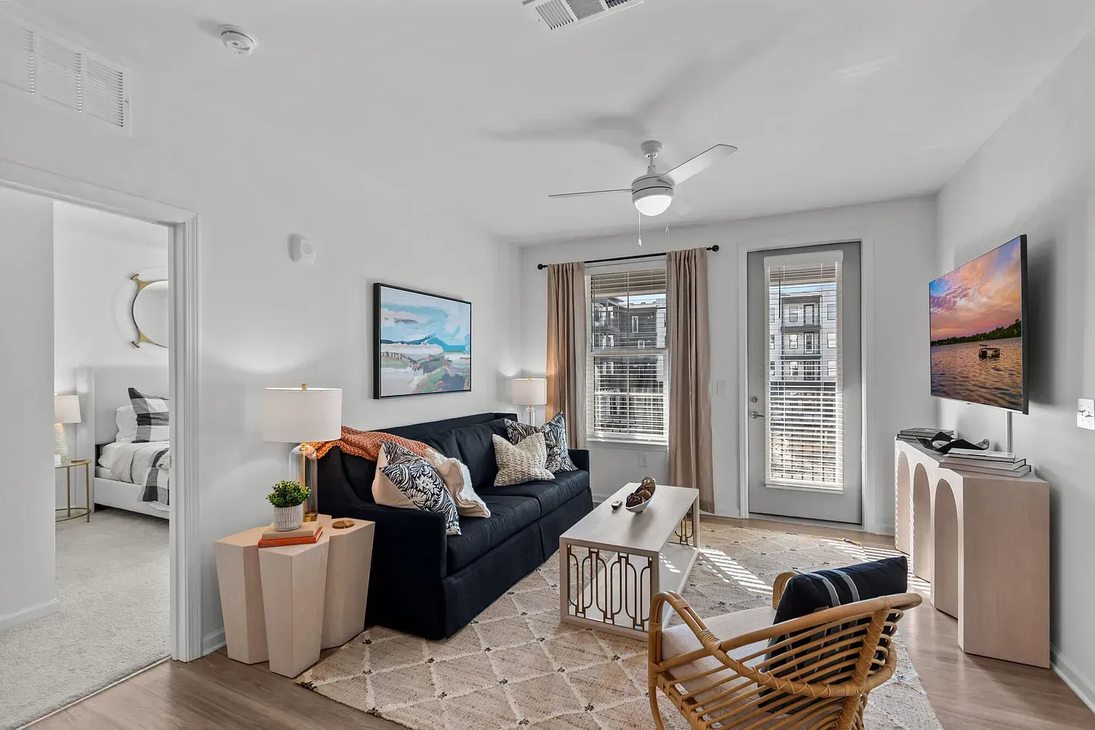Living room with navy sofa, coffee table, TV, ceiling fan, and balcony door; a glimpse of the adjacent bedroom.