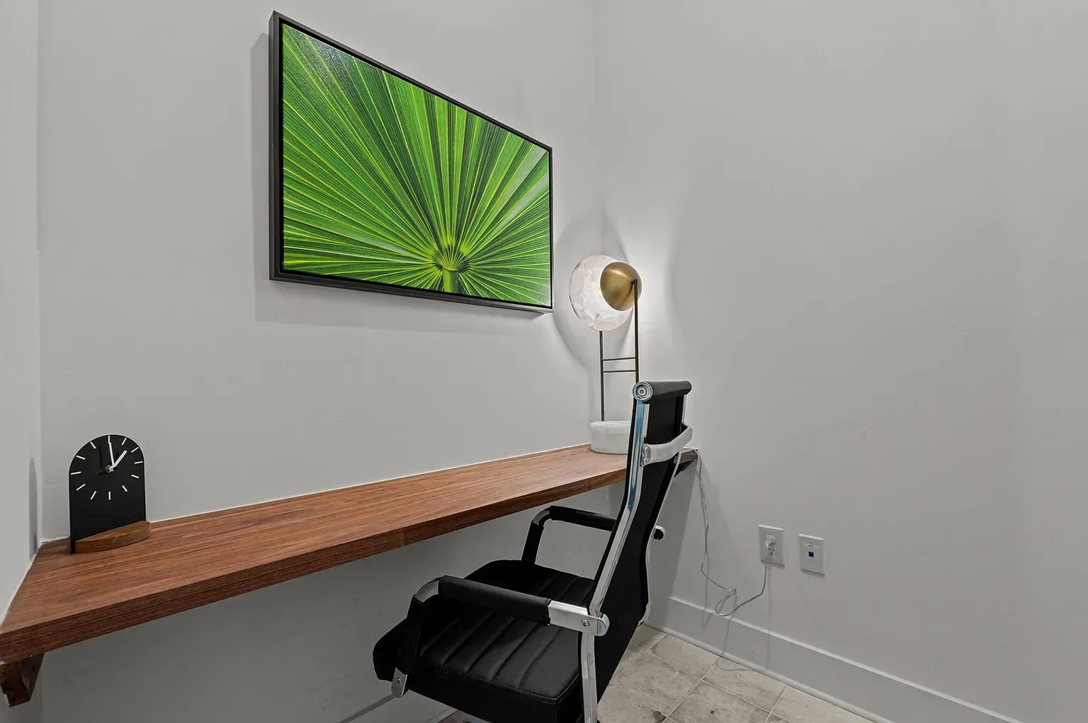 Home office corner with wooden desk, black office chair, wall lamp, and green abstract wall art.