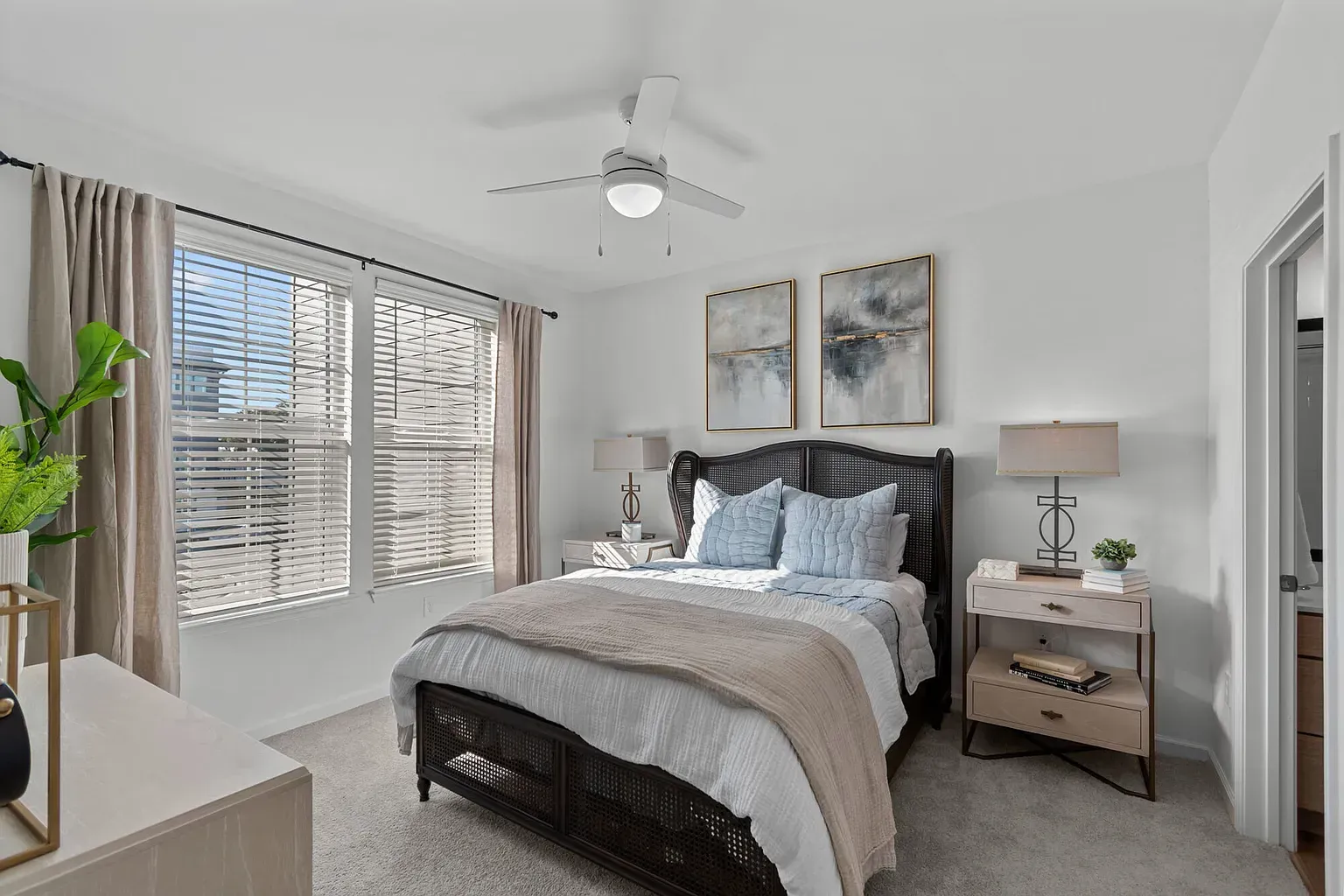 Bedroom in an apartment with a bed, two nightstands, lamps, and large windows with blinds.