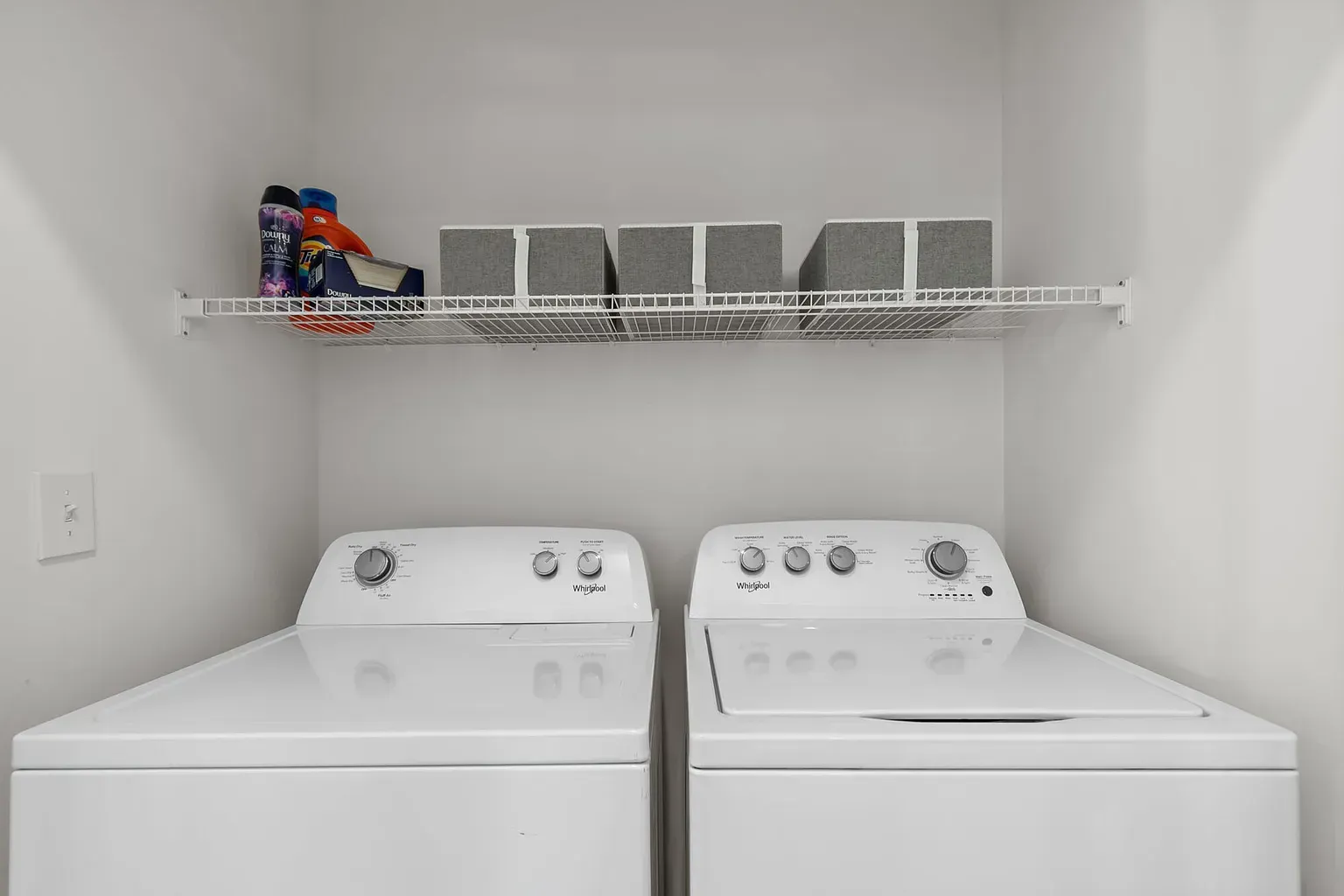 Two white top-loading washers in a small apartment laundry area with a wire shelf above.