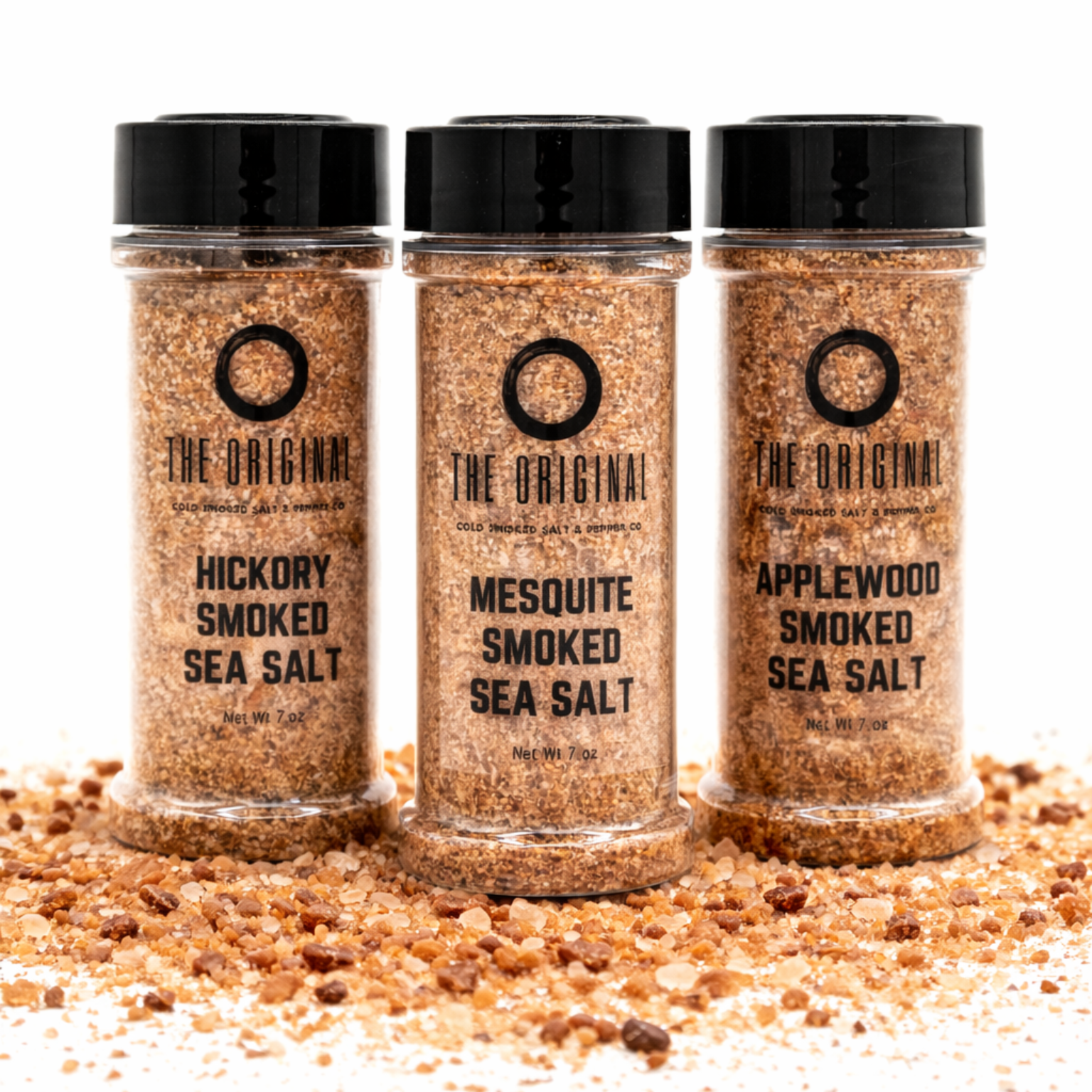 Three spice jars of smoked sea salt: hickory, mesquite, and applewood, on a bed of seasoning flakes