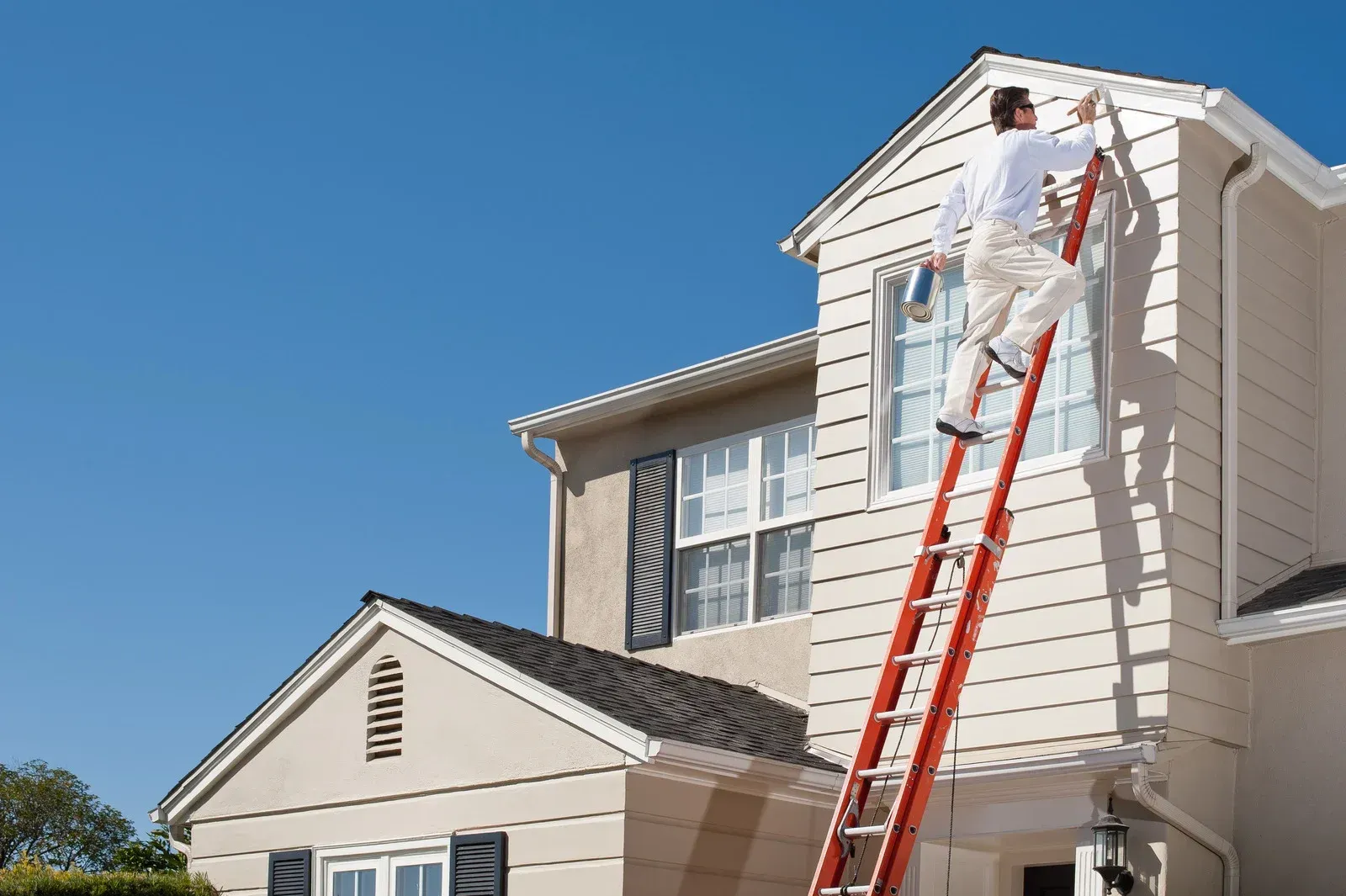 Worker on ladder painting the upper exterior of a house under a blue sky