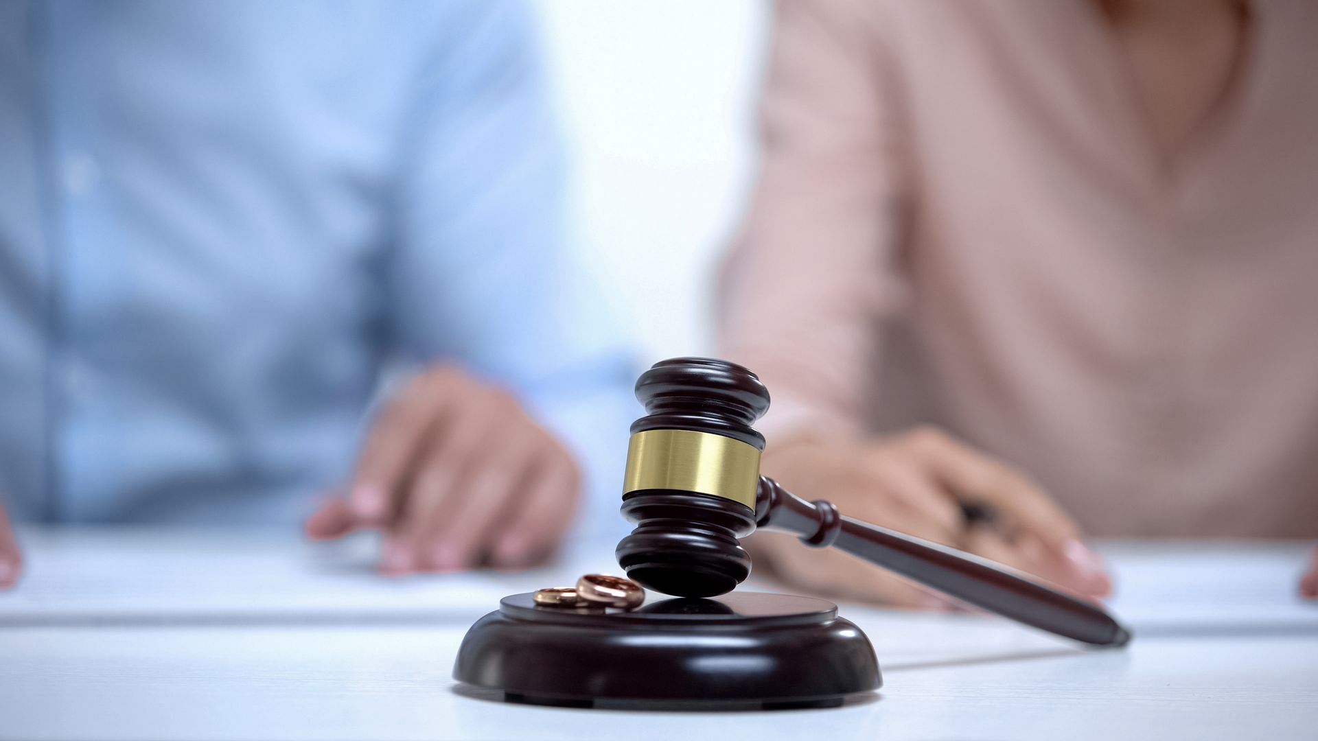 A judge’s gavel and two wedding rings on a desk as a couple sits across the table. A judge’s gavel and two wedding rings on a desk as a couple sits across the table.