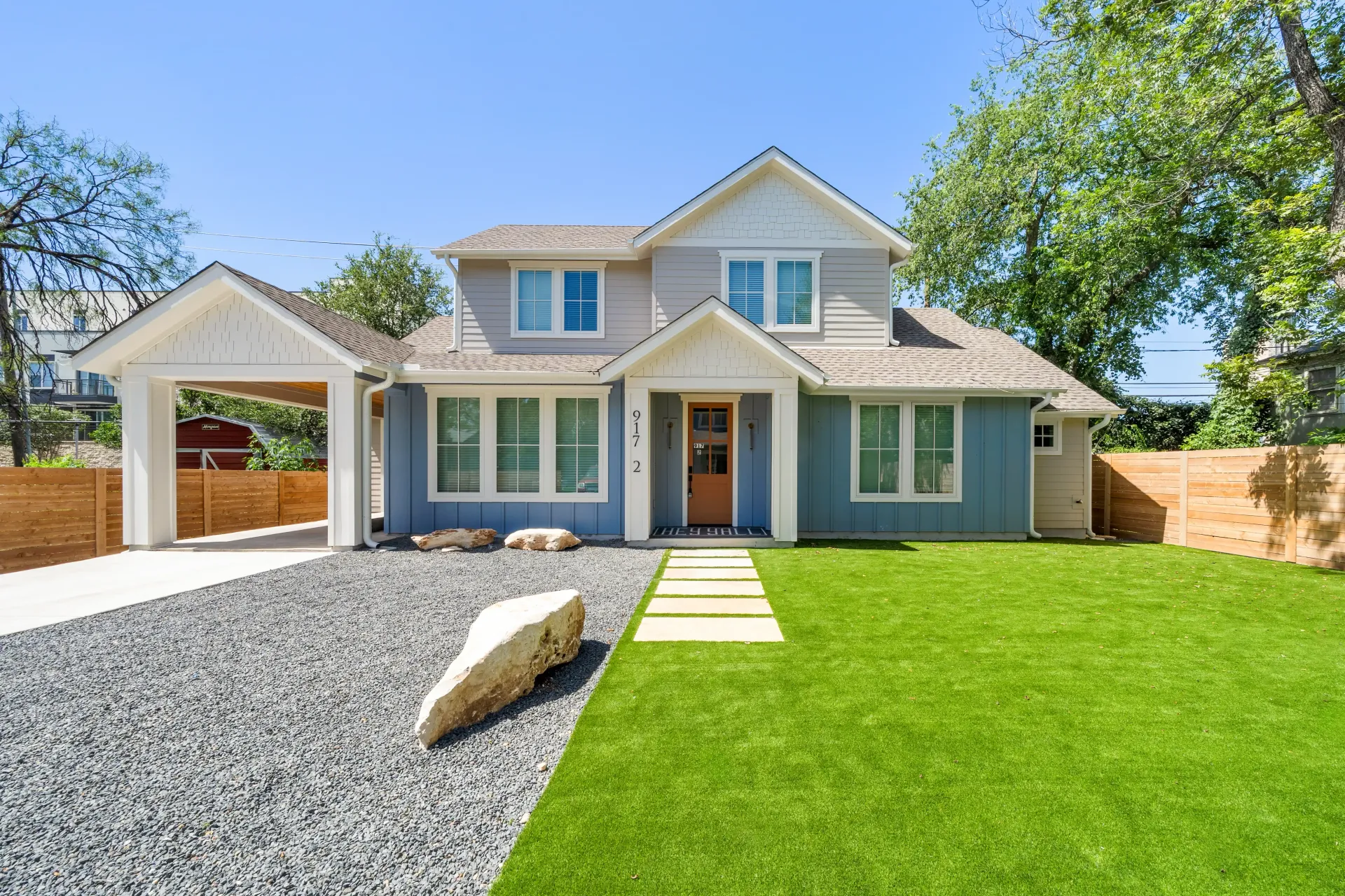 Two-story house with blue siding, a carport, and green lawn; gray gravel path, sunny day.