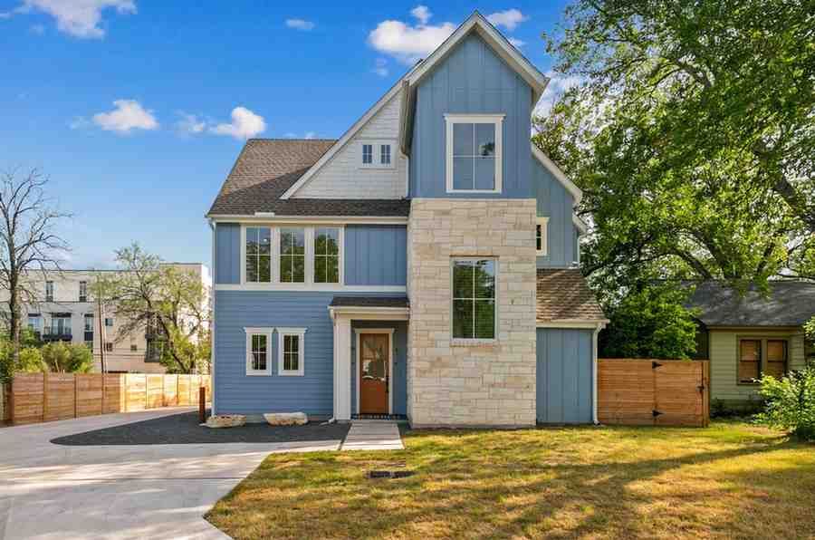 Two-story blue house with stone accents, brown door, and brown roof. Front yard with driveway and fence.