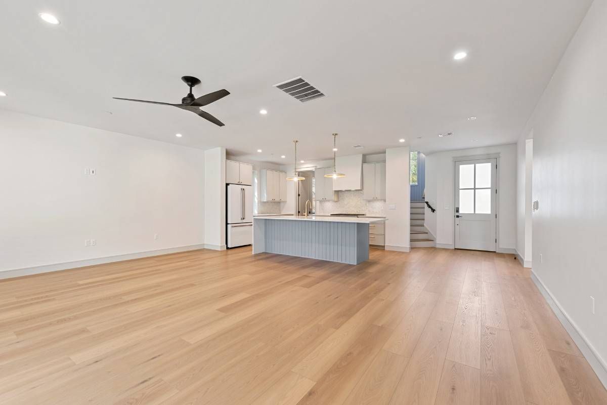Spacious interior: light wood floors, white walls, kitchen island, refrigerator, ceiling fan, and door.