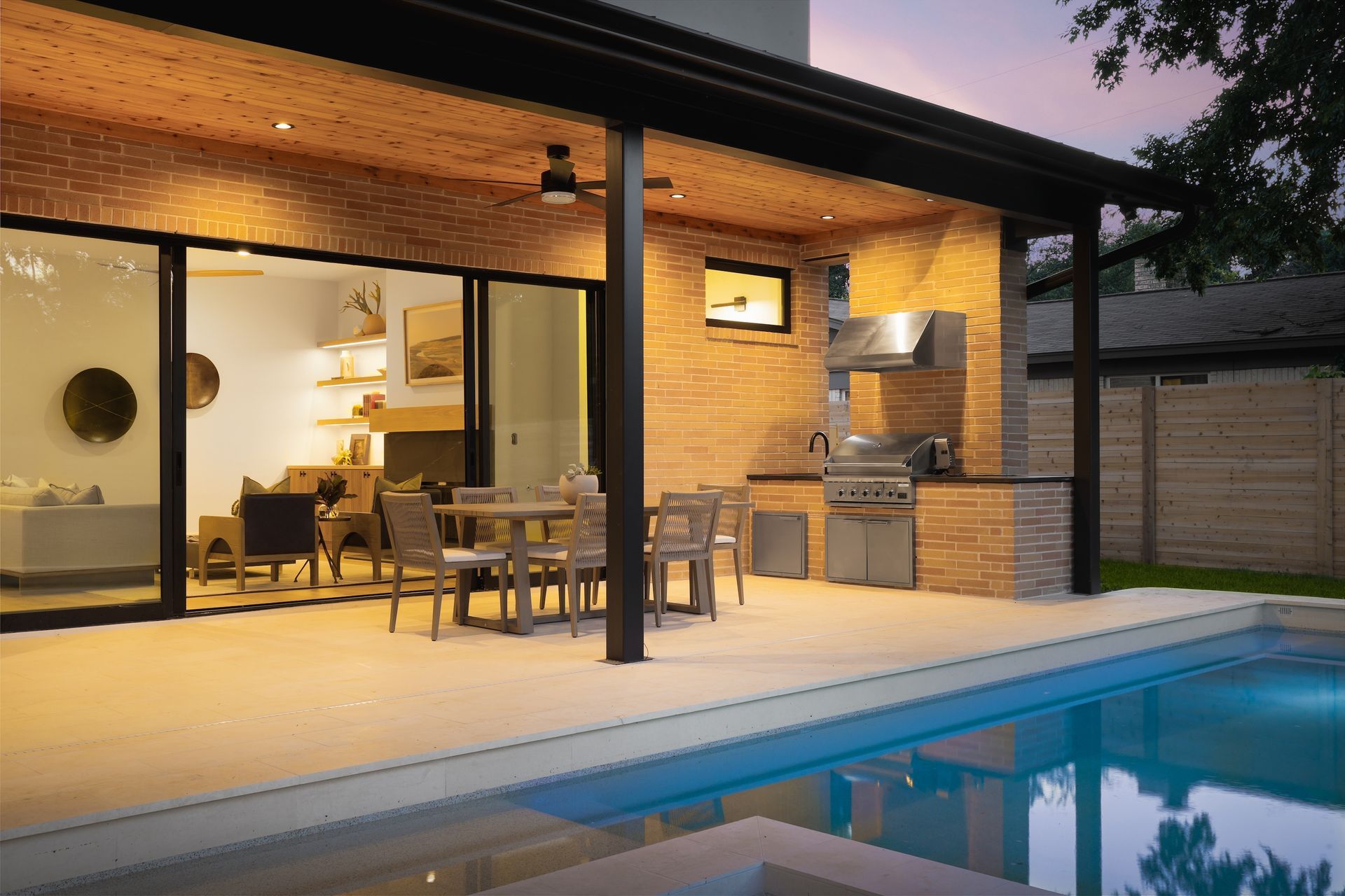 Outdoor patio with a pool, kitchen, and dining area. Warm lighting and a clear night sky.