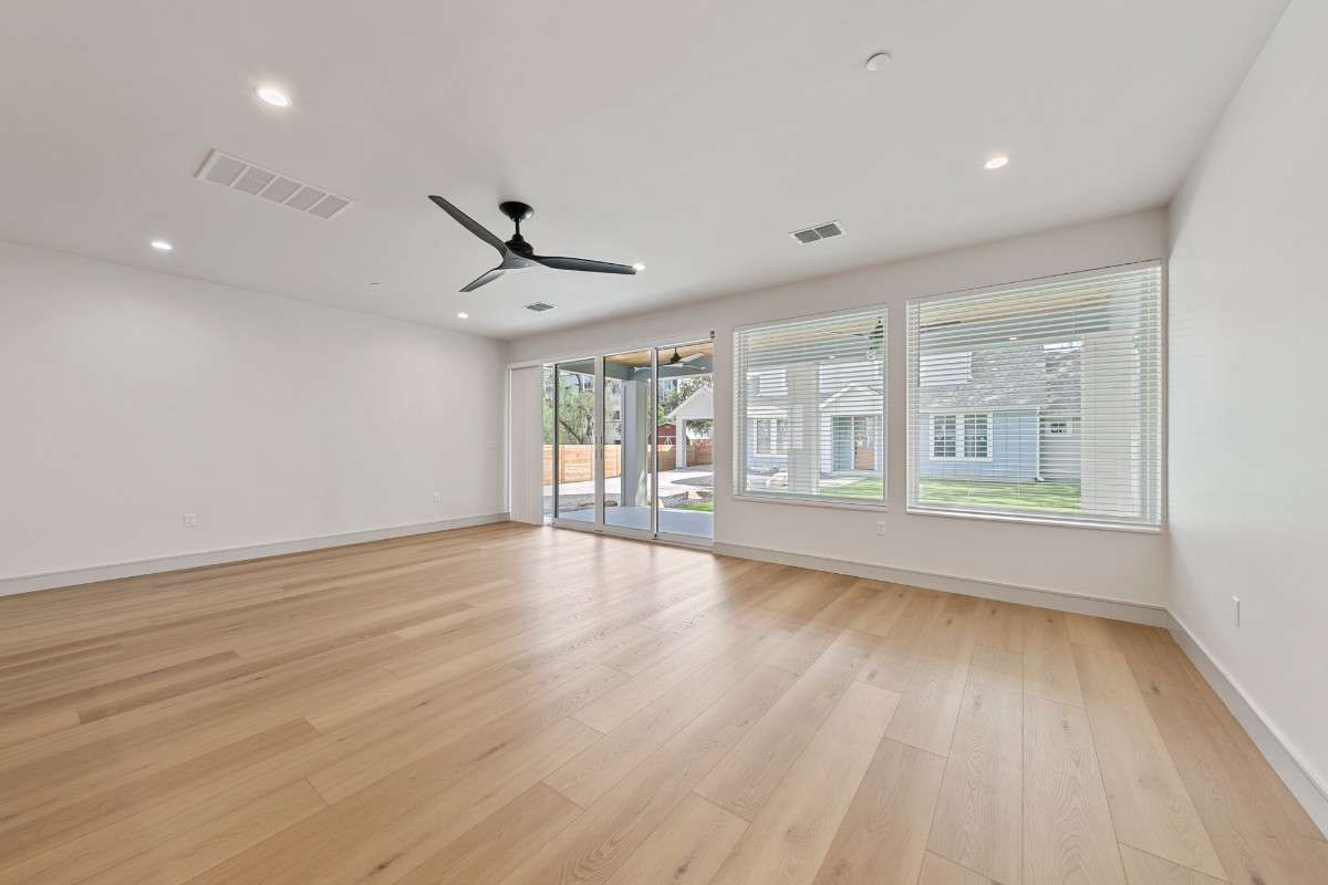 Empty room with hardwood floors, white walls, and a ceiling fan, with large windows looking outside.