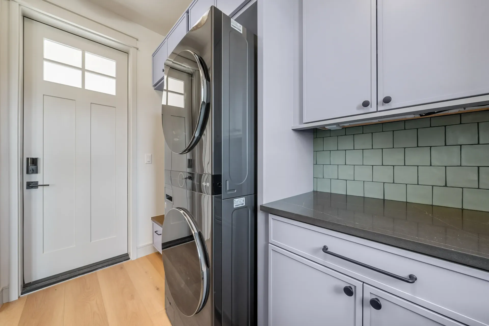 Laundry room with stacked silver washer/dryer, white door, gray cabinets, and a gray countertop.