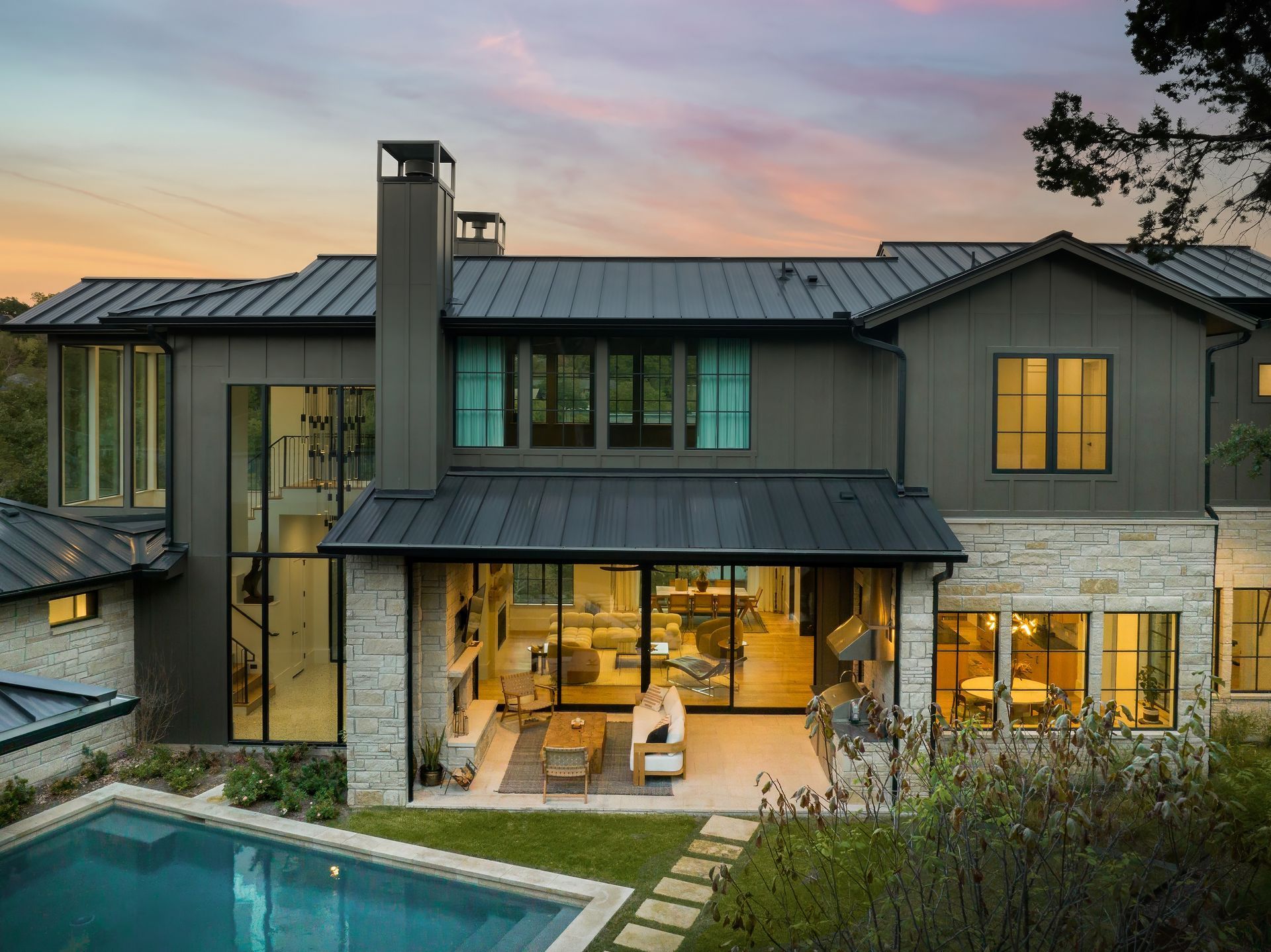 Luxury home exterior with pool. Stone and dark gray siding. Evening sky. Large glass doors open to the living area.