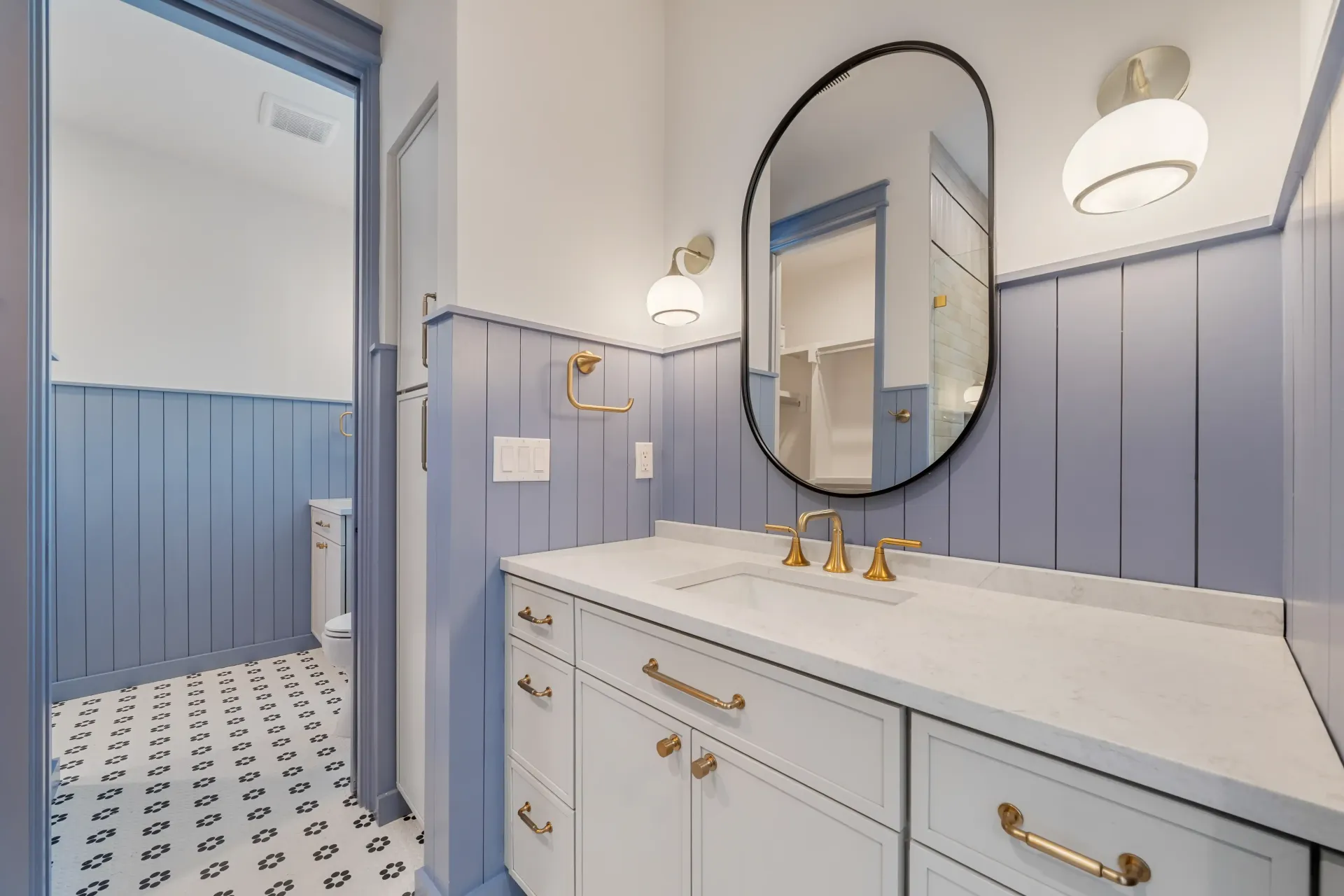 Bathroom with blue and white paneling, white vanity with gold hardware, oval mirror, and patterned floor.