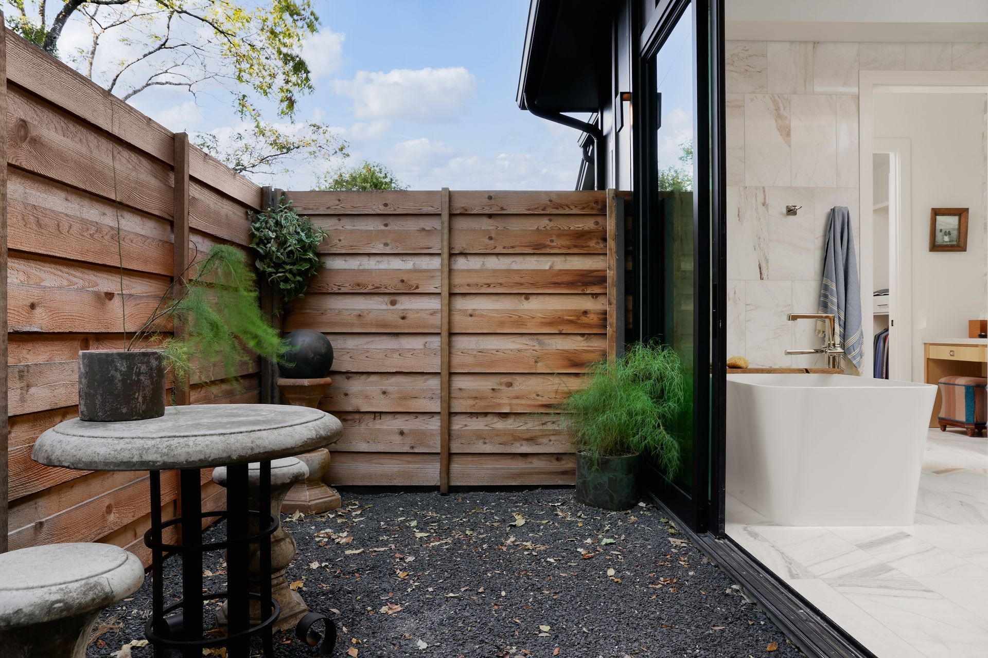 Outdoor patio with wooden fence, round stone table, plants, and view into a modern bathroom.