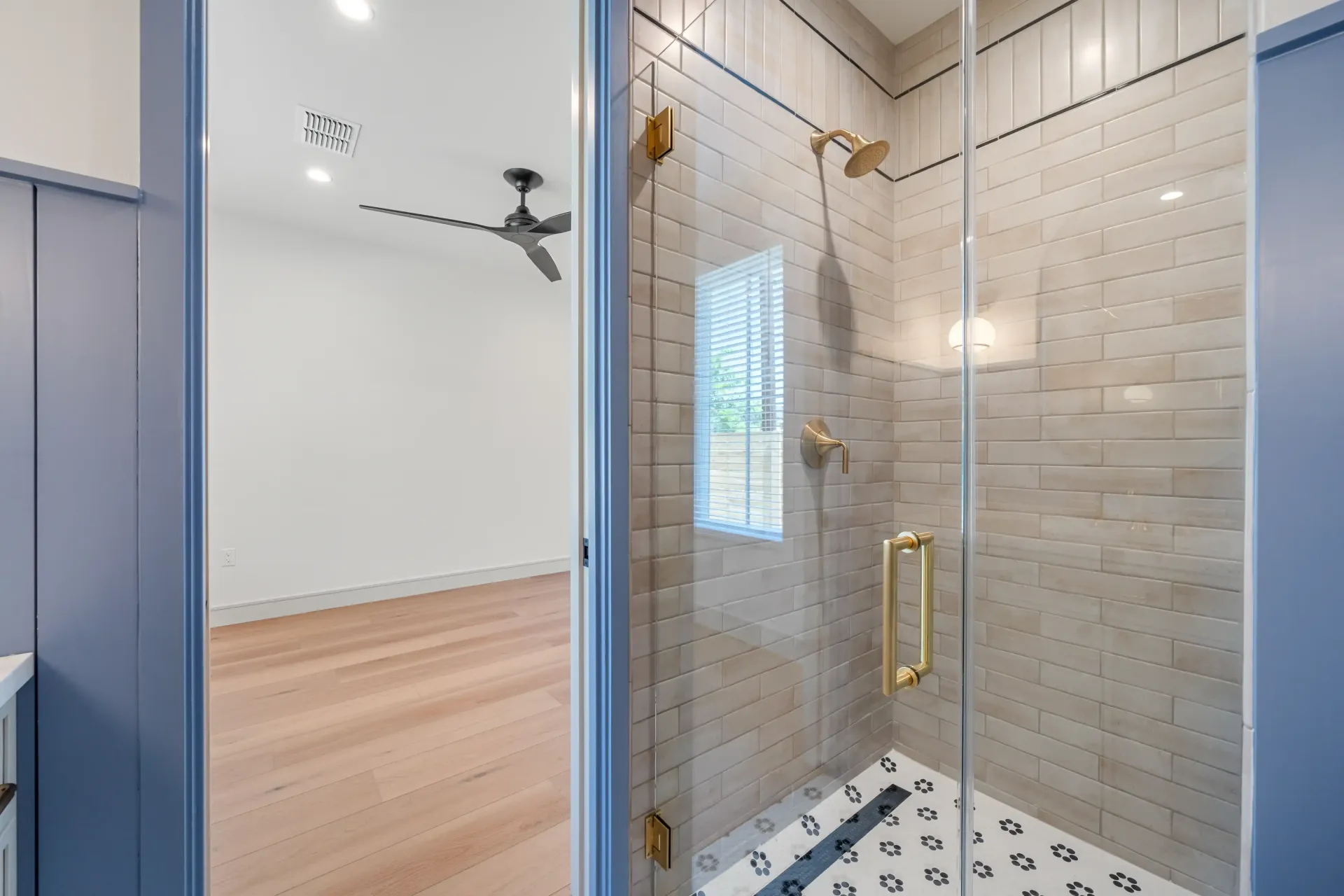 Shower with glass door, gold fixtures, and patterned floor. Open door reveals bedroom with fan.