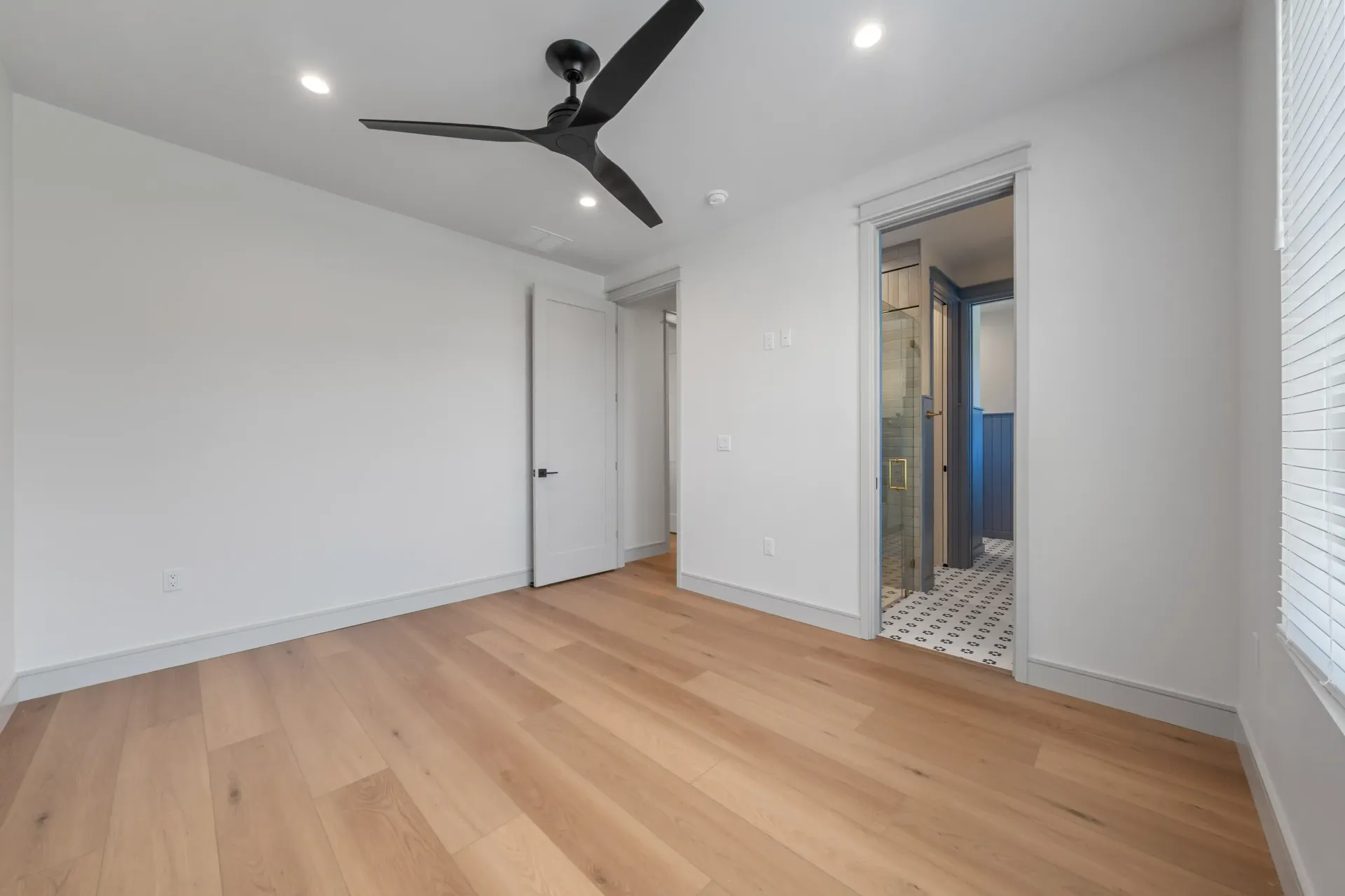 Empty bedroom with light wood flooring, white walls, black ceiling fan, and open bathroom doorway.