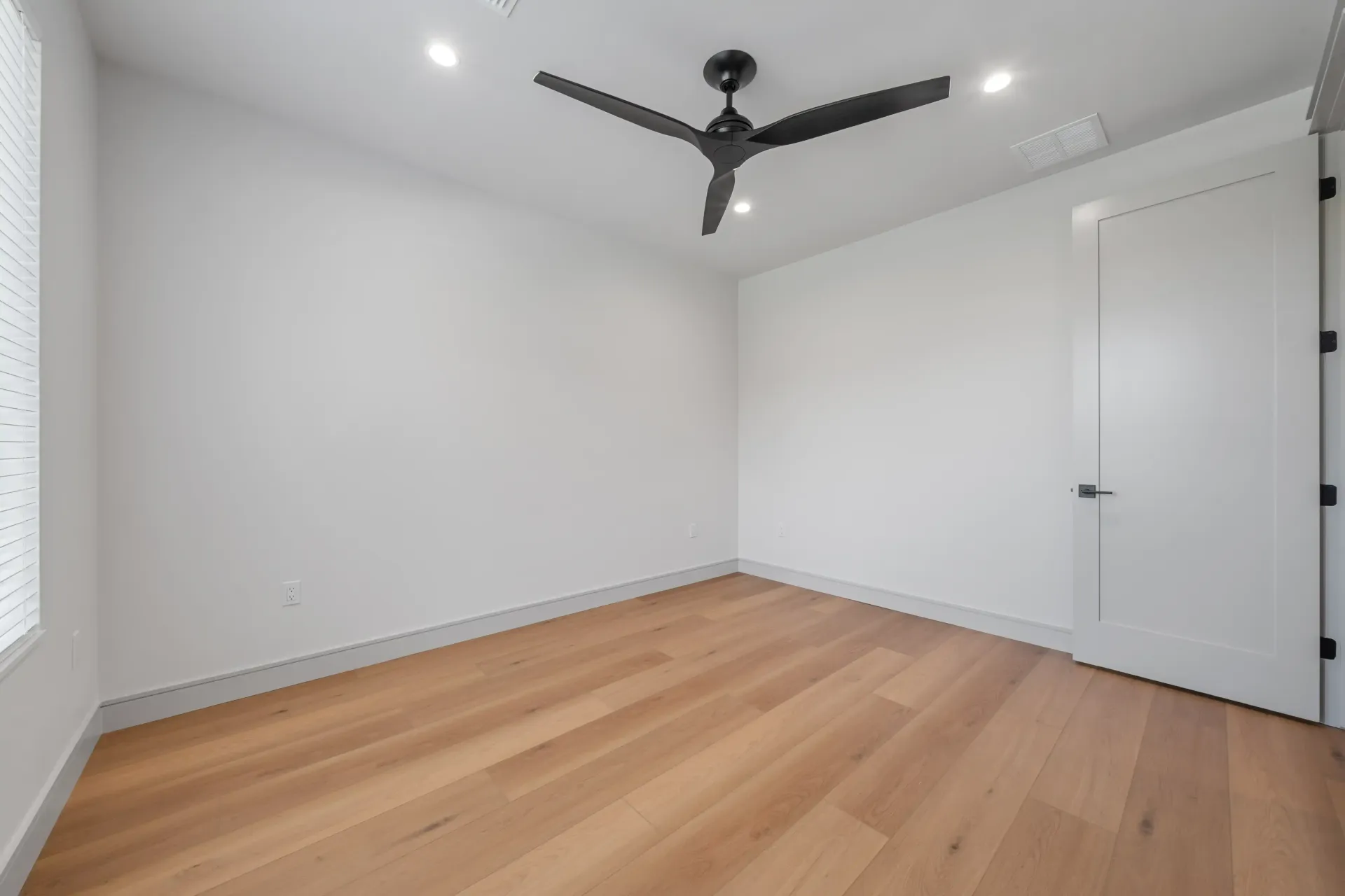 Empty room with light wood floors, white walls, black ceiling fan, and a closed door.