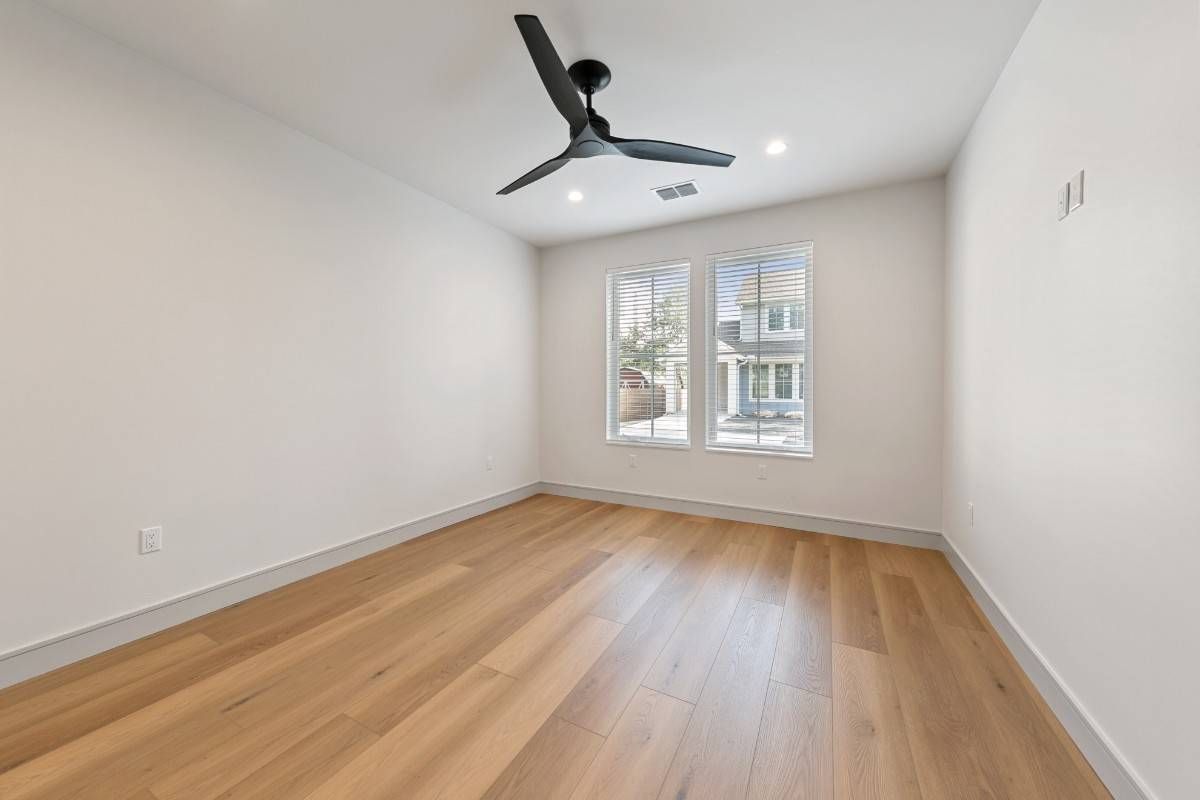 Empty room with hardwood floors, white walls, ceiling fan, and window looking out onto a house.
