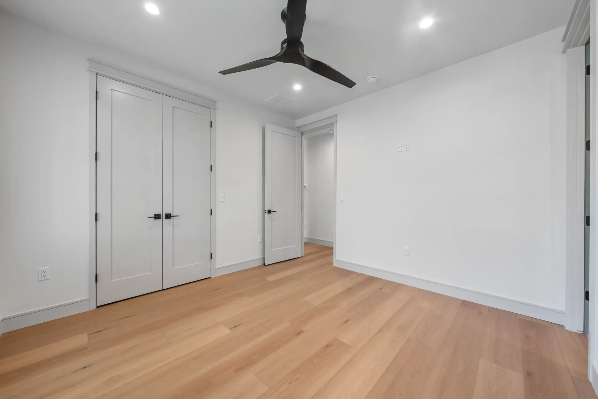 Empty room with white walls, wood floors, and black ceiling fan. Two white doors and a doorway.