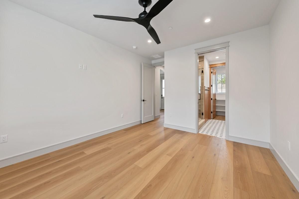 Empty room with light wood floor, white walls, and a black ceiling fan; doorway to a bathroom.