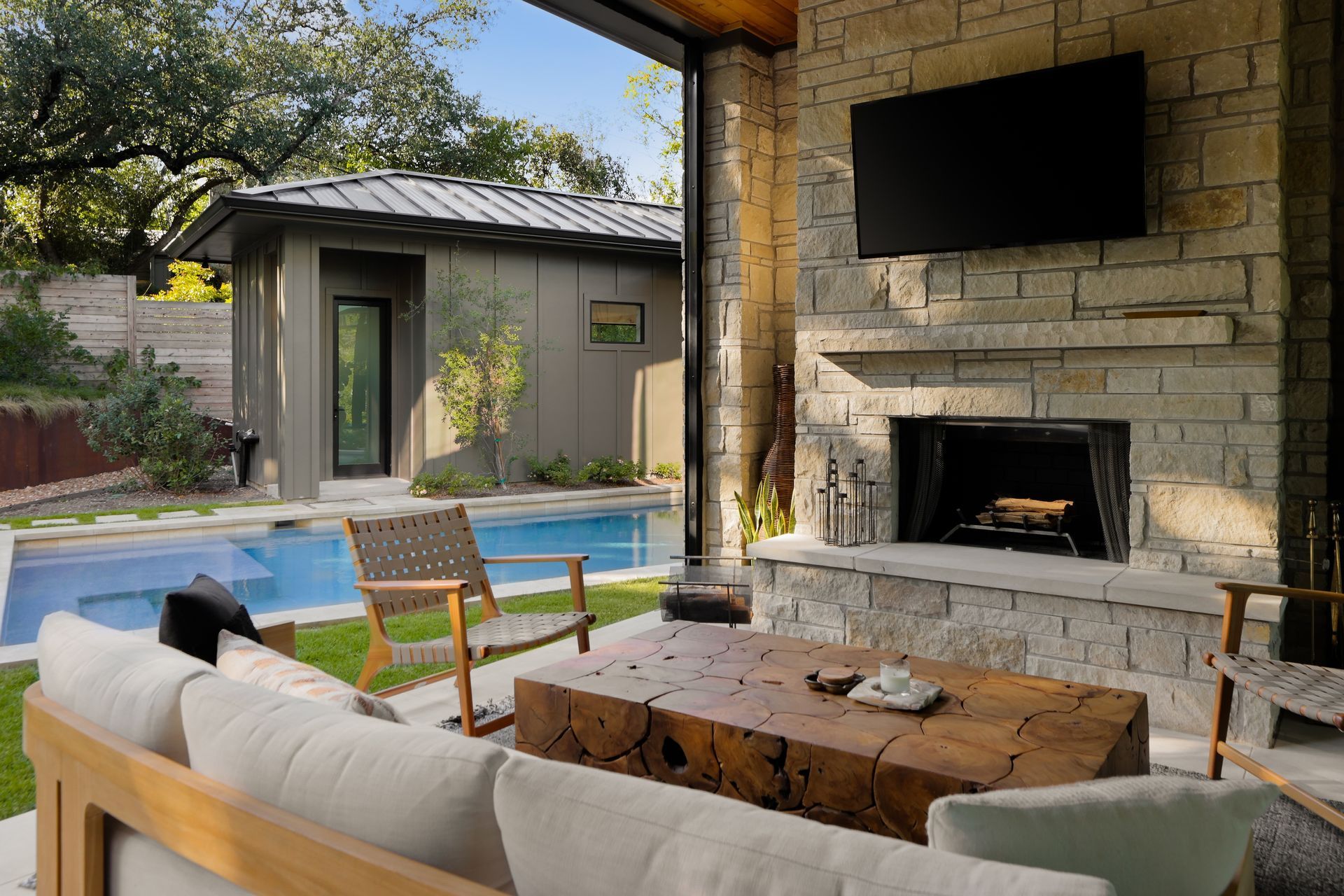 Outdoor living space with a pool. Stone fireplace and TV, seating area, and a building in the background.