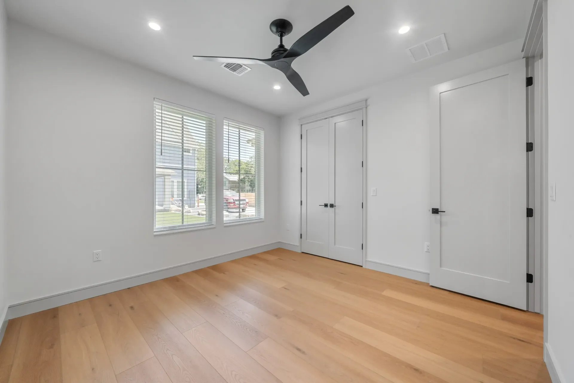 Empty room with wood floor, white walls, two windows, two doors, and a ceiling fan.