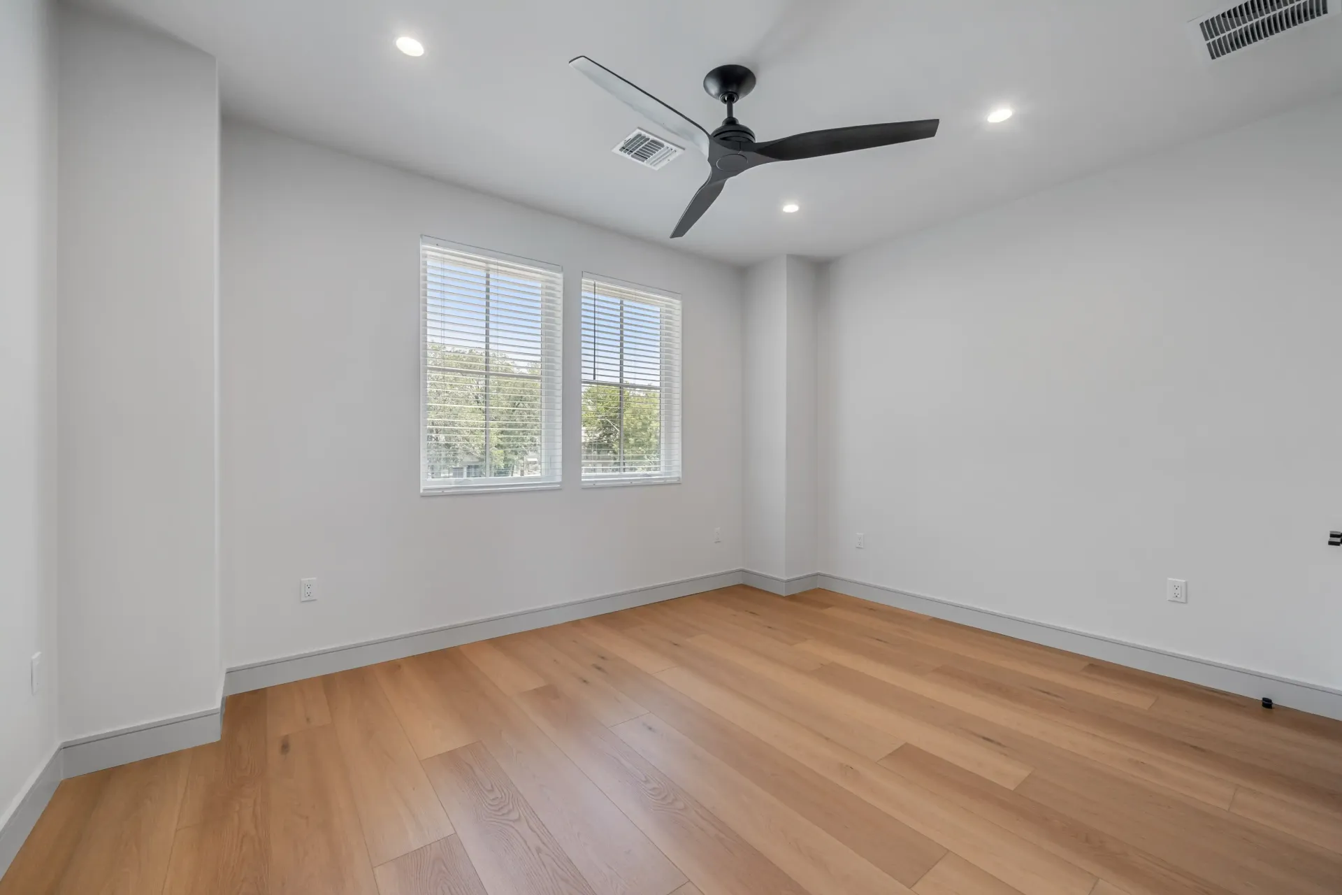 Empty room with light wood floors, two windows, ceiling fan, and white walls.