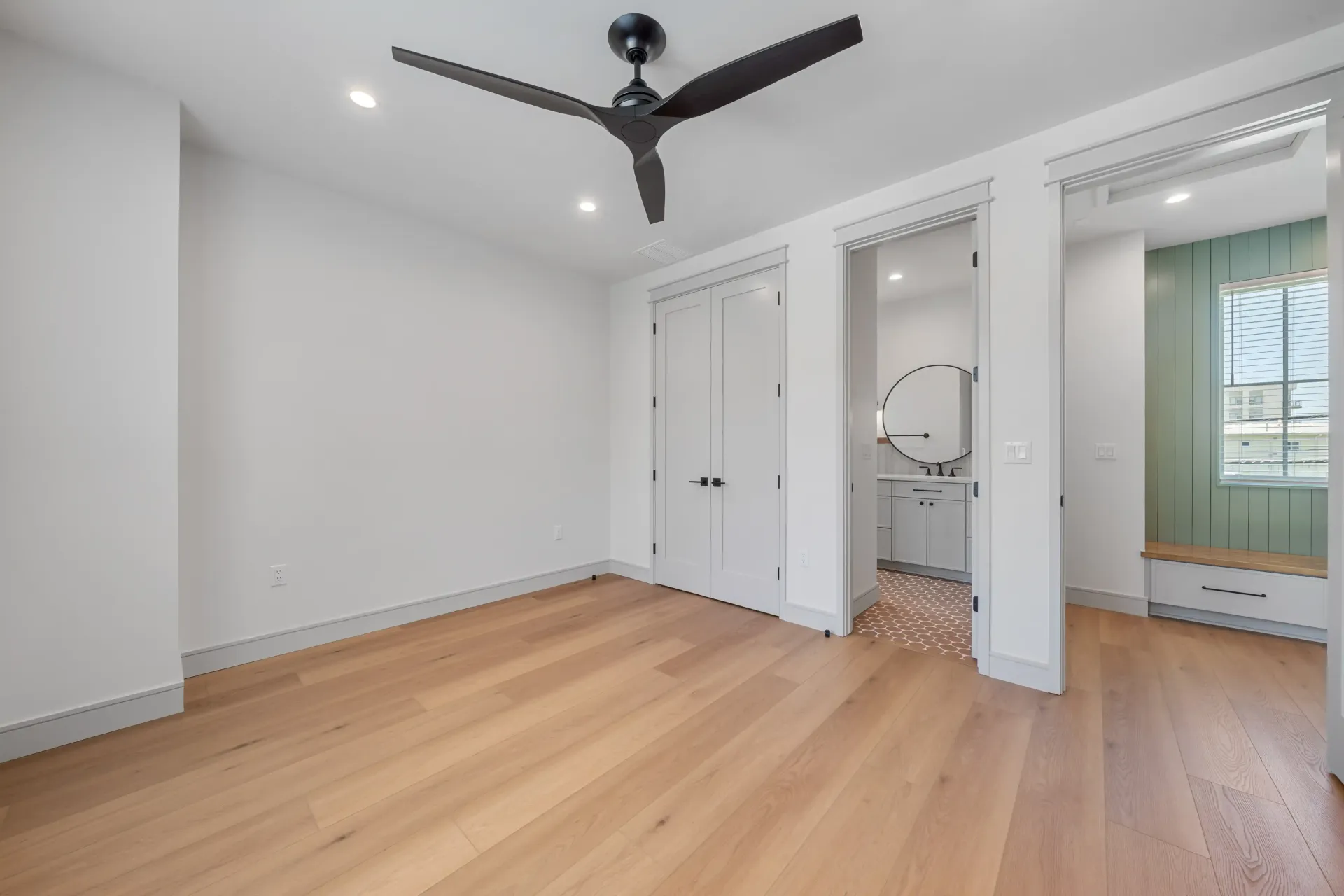 Empty room with wood floor, white walls, black ceiling fan, and doors leading to a bathroom.