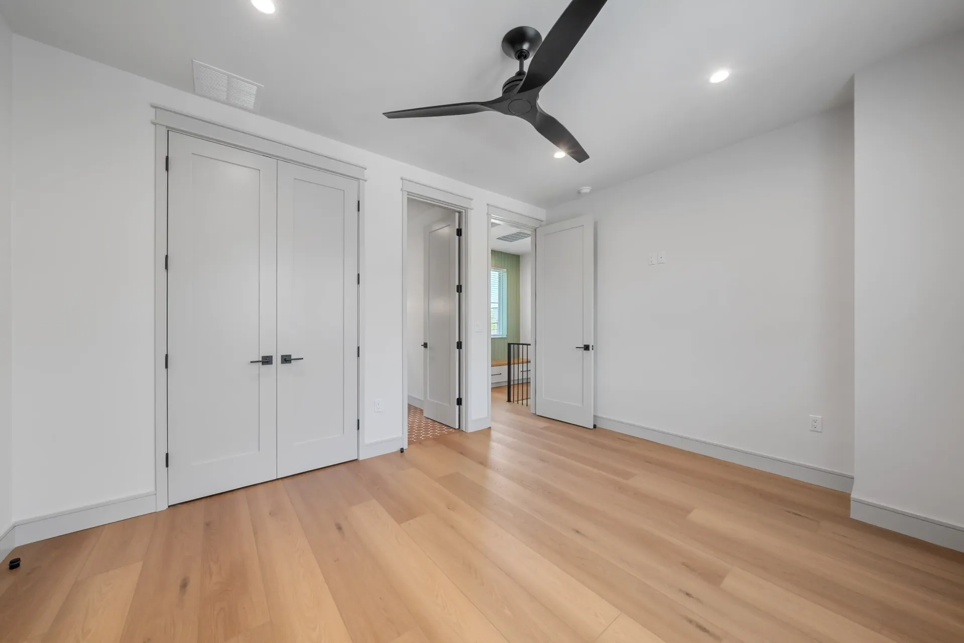 Empty room with light wood floors, white walls, and black ceiling fan. Three white doors.