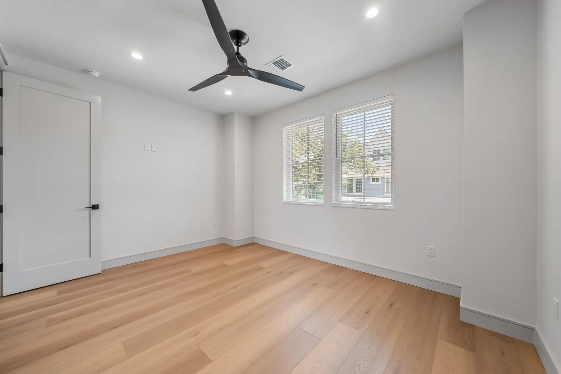 Empty room with wood floor, white walls, two windows, ceiling fan.