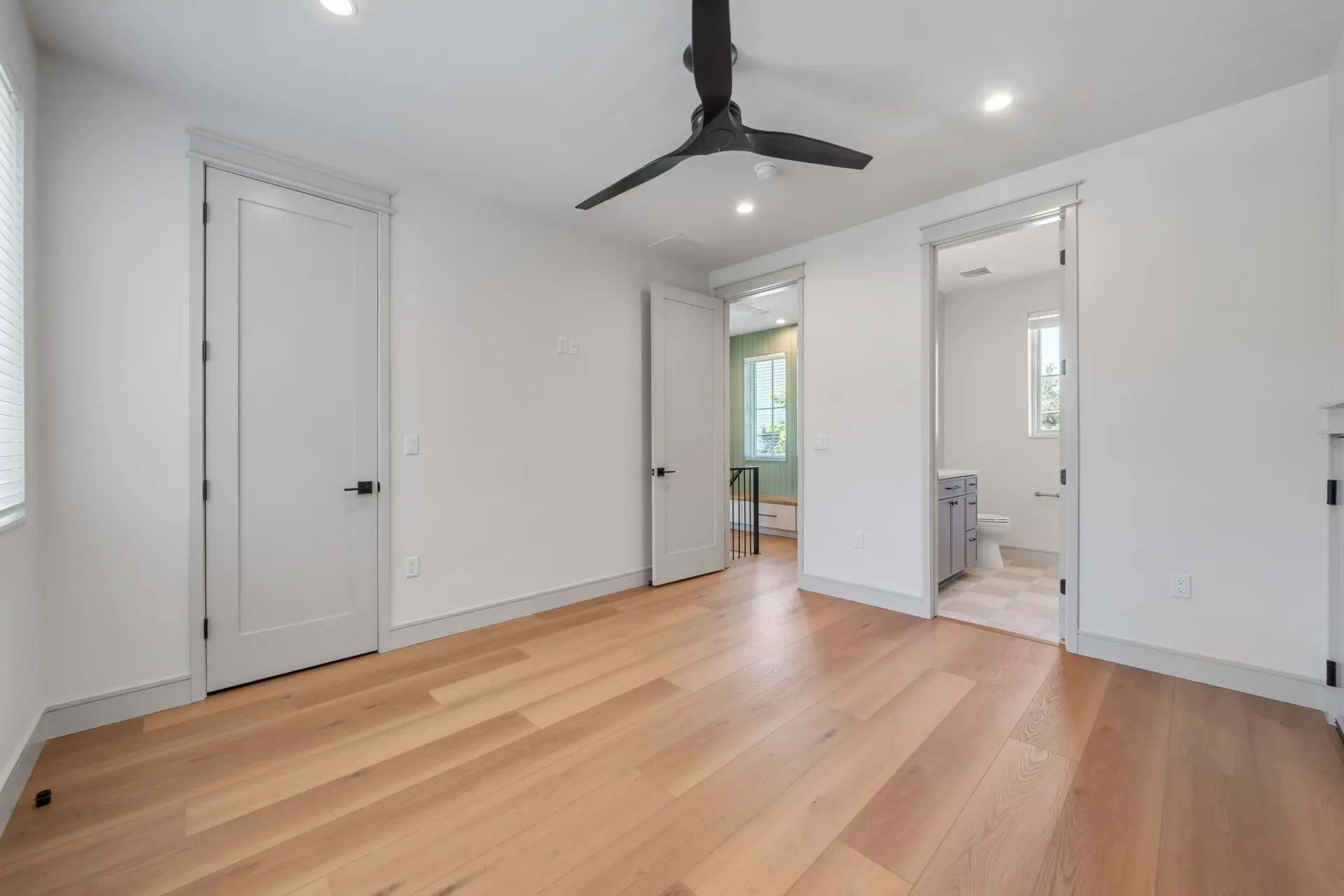 Empty bedroom with hardwood floors, white walls, and a black ceiling fan; doors lead to bathroom and other rooms.