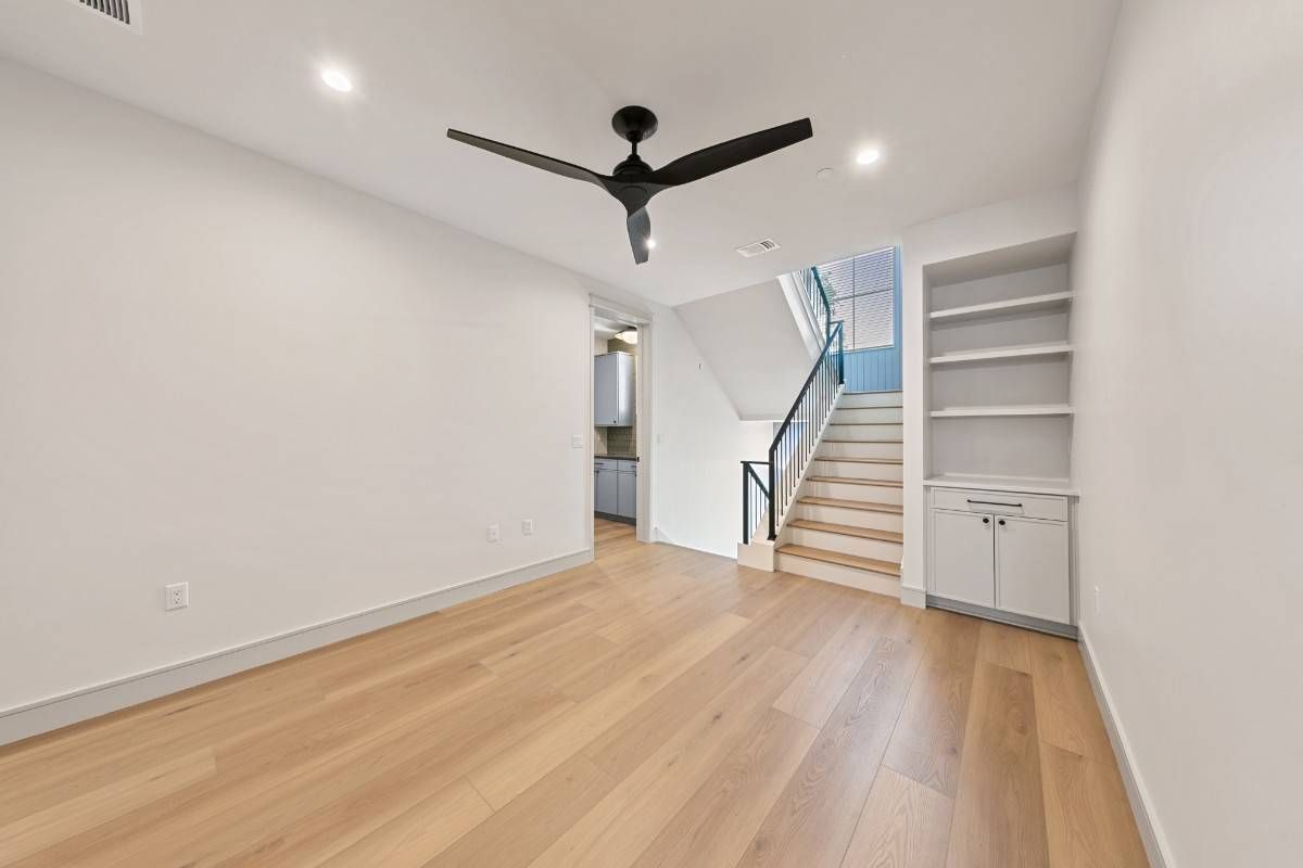 Empty room with wood floors, built-in shelves, and a staircase leading upwards. Black ceiling fan.