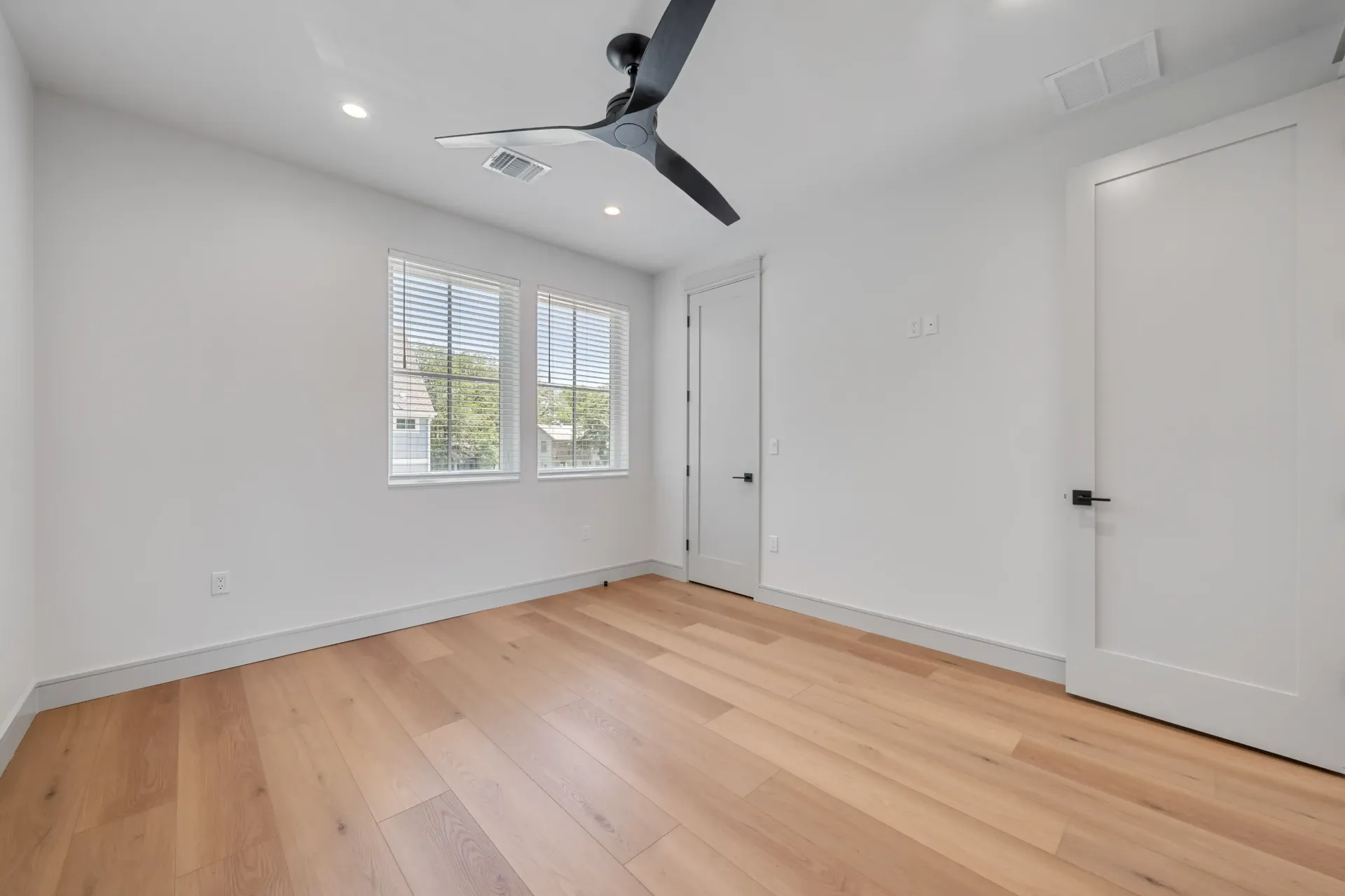 Empty room with light wood floor, white walls, two white doors, window, black ceiling fan.