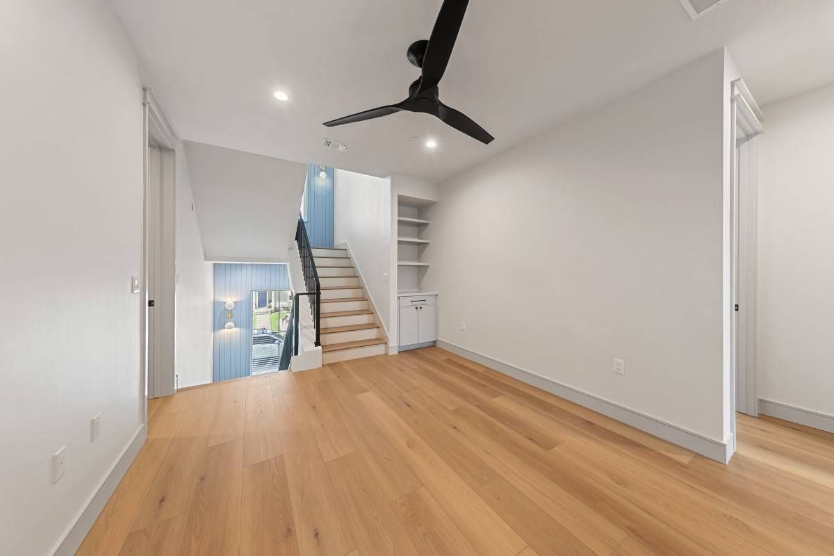 Empty room with wood floors, stairs, and ceiling fan. White walls, built-in shelving.