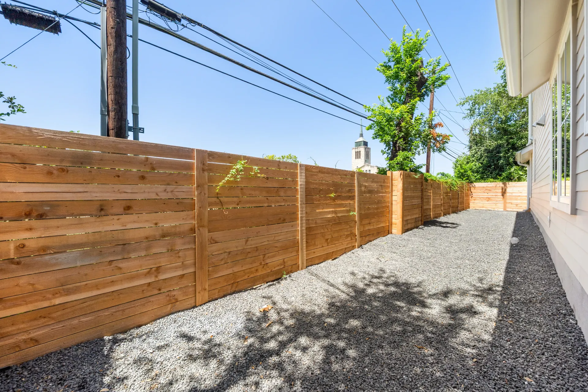 Wooden fence beside gravel pathway under a blue sky, with utility pole and trees in the background.