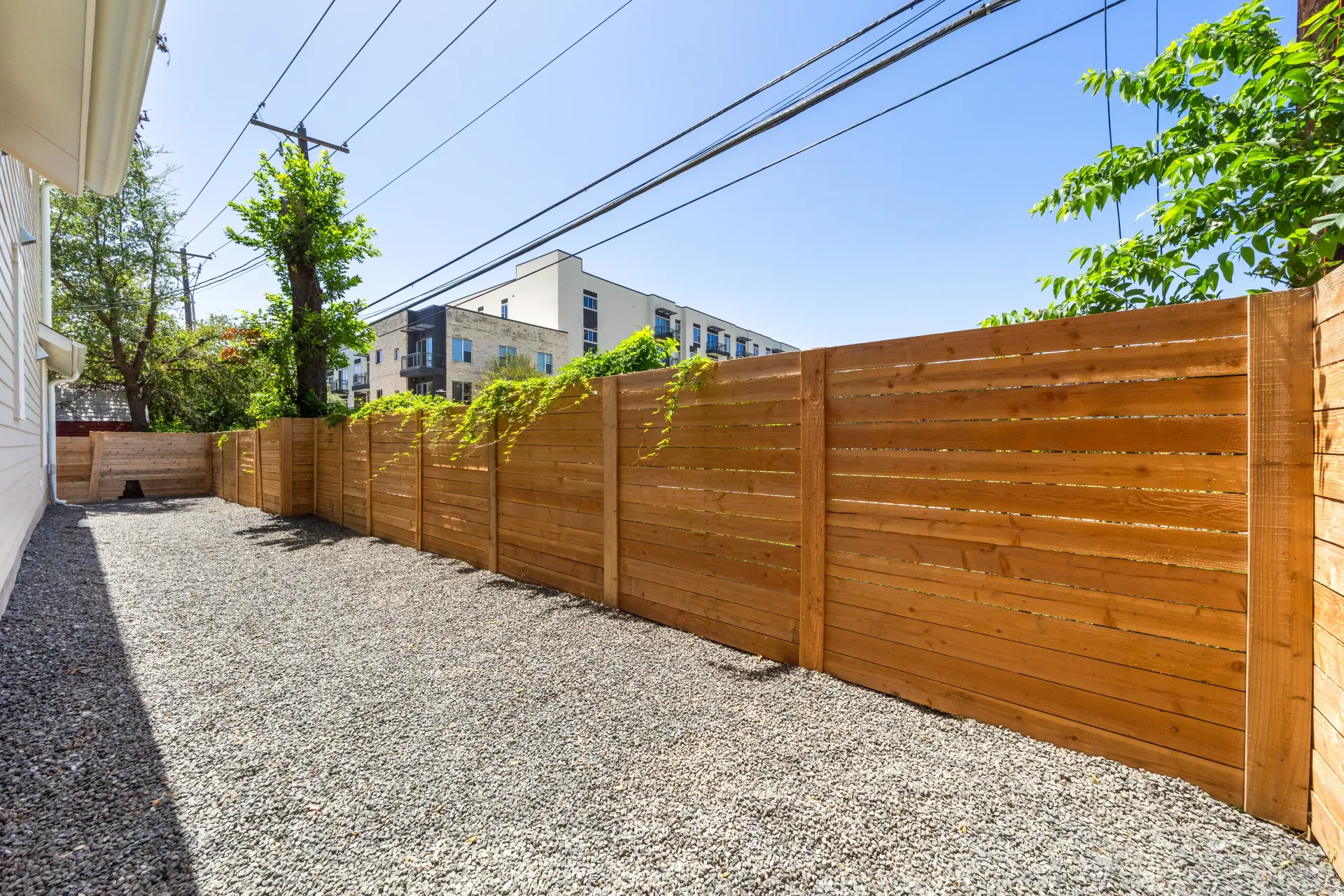 Gravel backyard with a brown wooden fence, and overhead power lines under a bright blue sky.