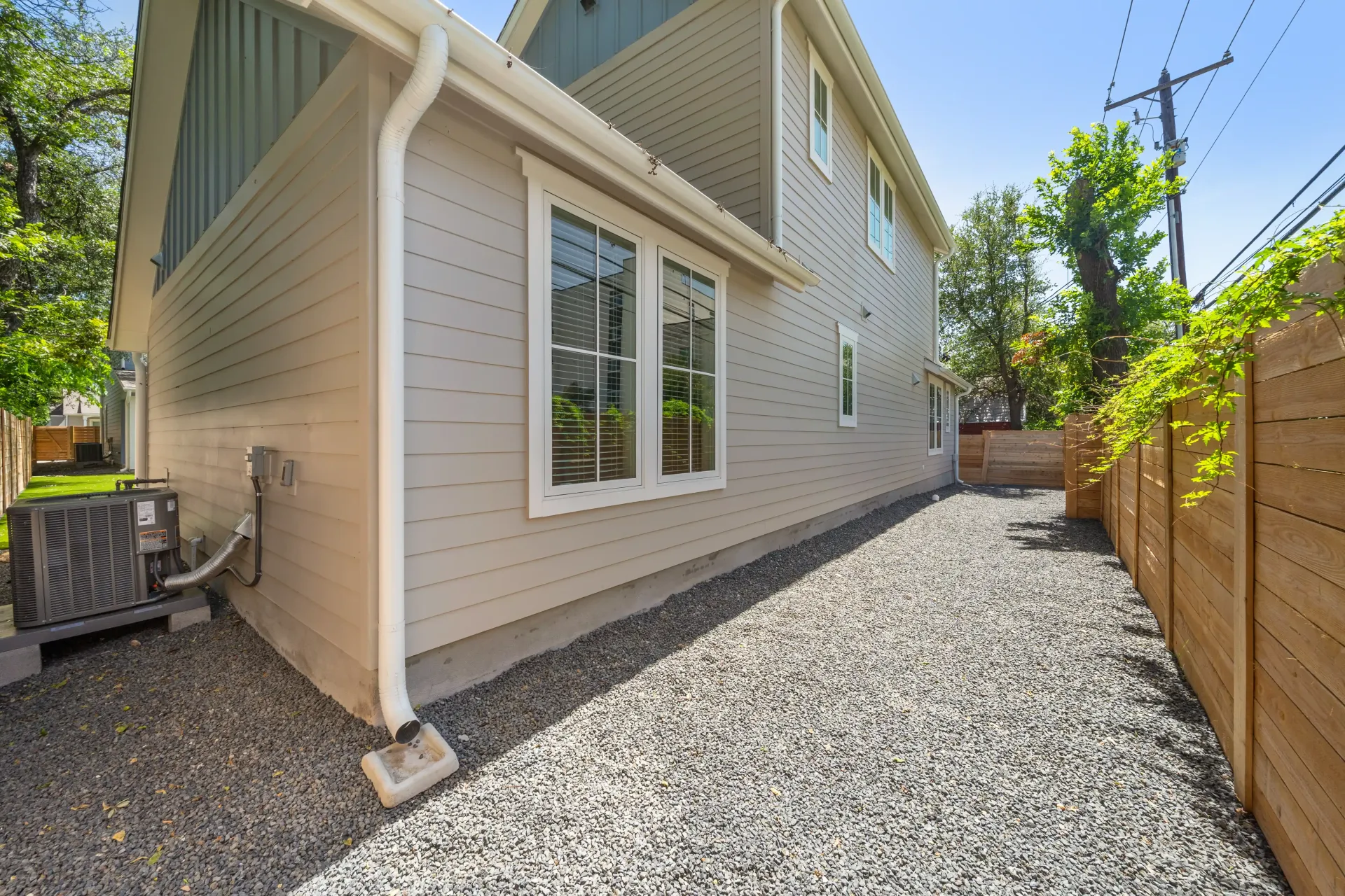 Side view of a two-story beige house with gravel pathway, wooden fence, and air conditioning unit.