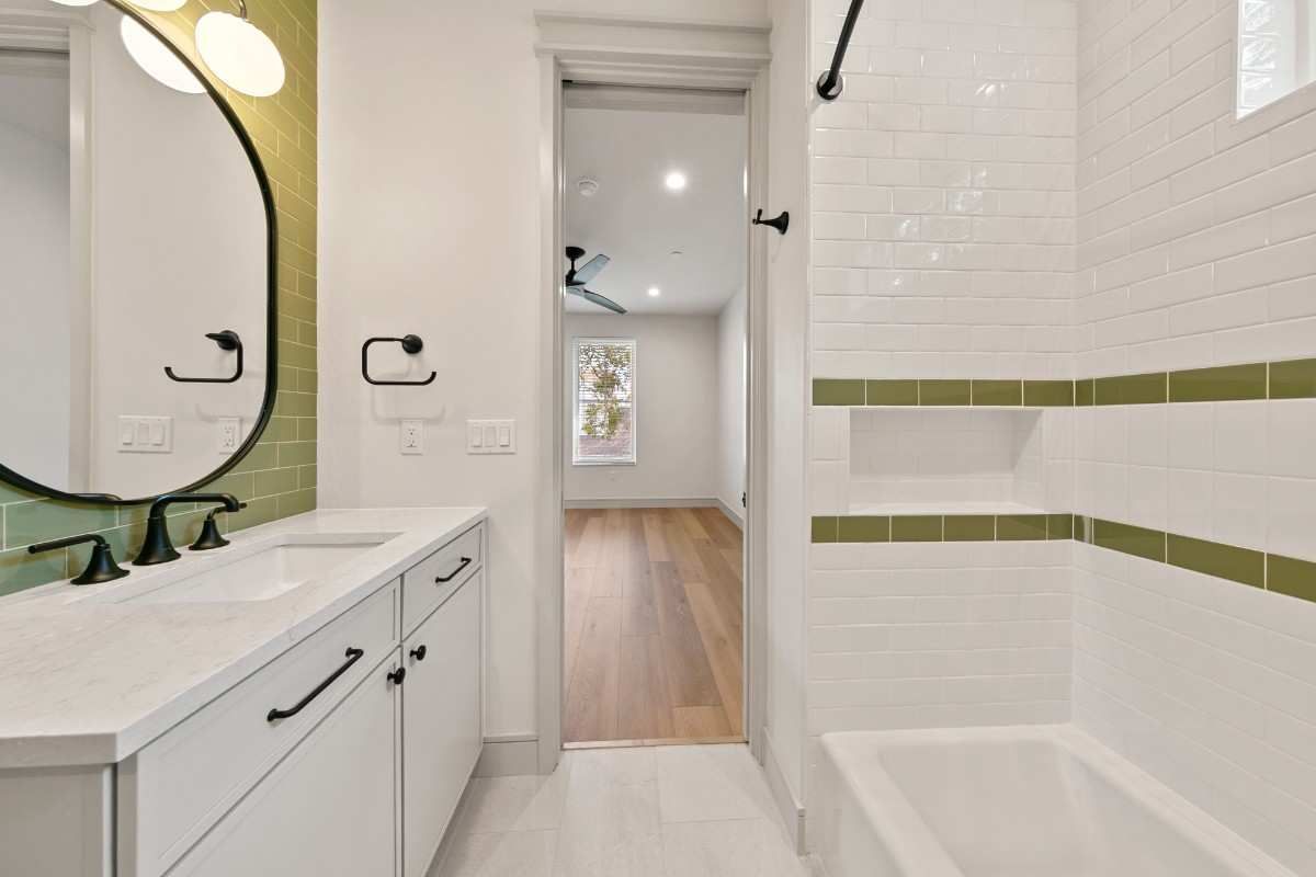 Bathroom with white vanity, green accent tiles, and a view into a room with wood floors.