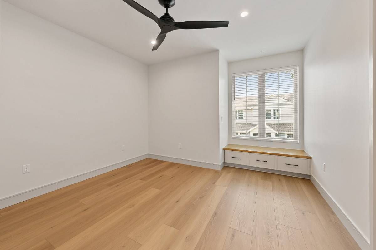 Empty room with hardwood floors, white walls, and a window with a built-in bench. Black ceiling fan.