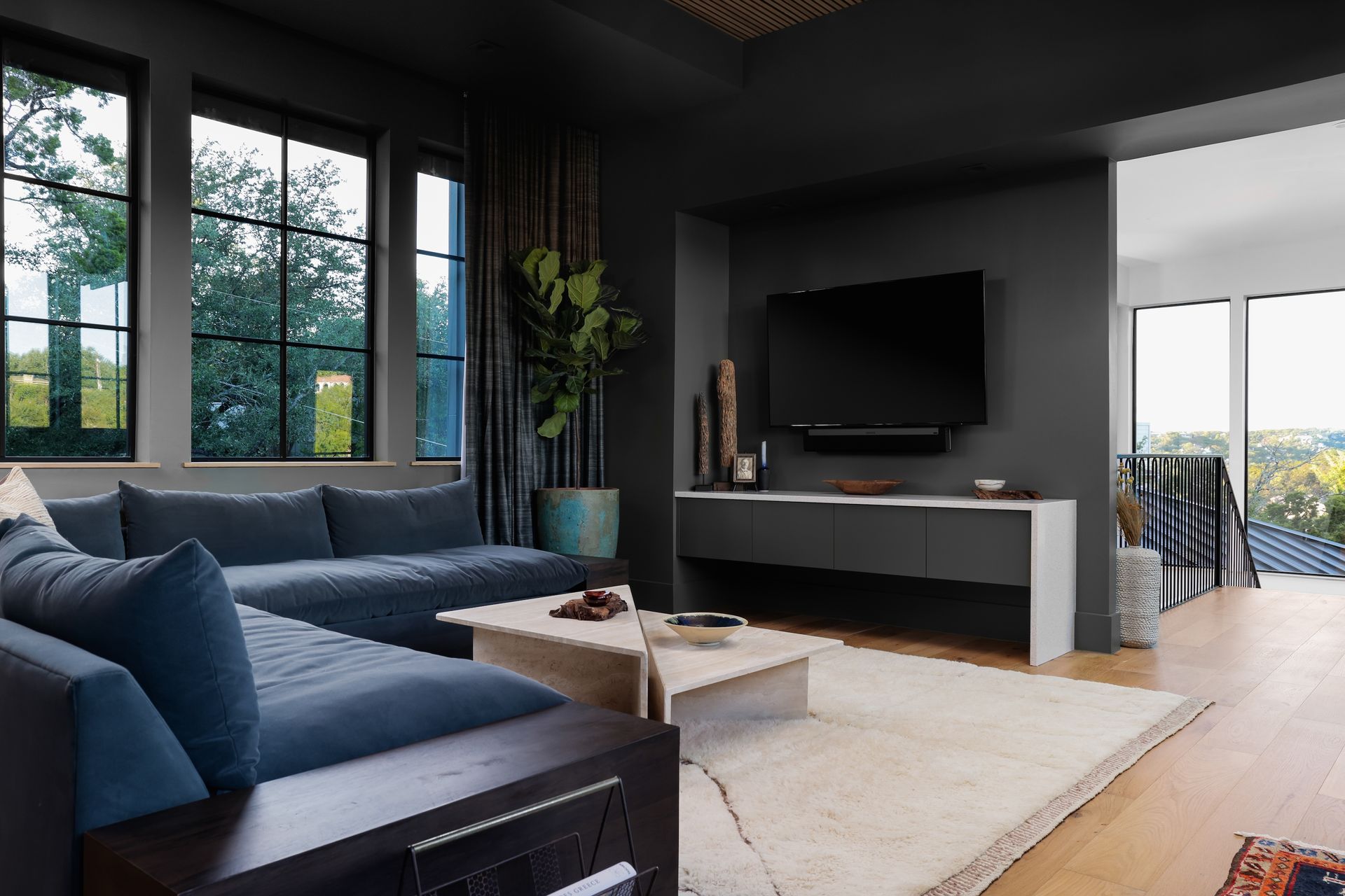 Dark living room with blue sectional sofa, TV, and large windows overlooking trees.