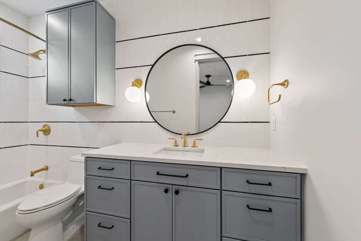 Modern bathroom with gray vanity, round mirror, and gold fixtures.