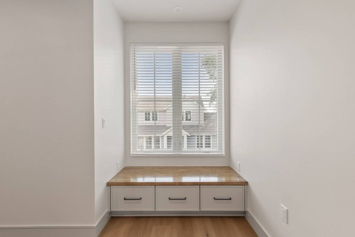 Built-in window seat with drawers, window with blinds overlooking a building. White walls, wood floor.