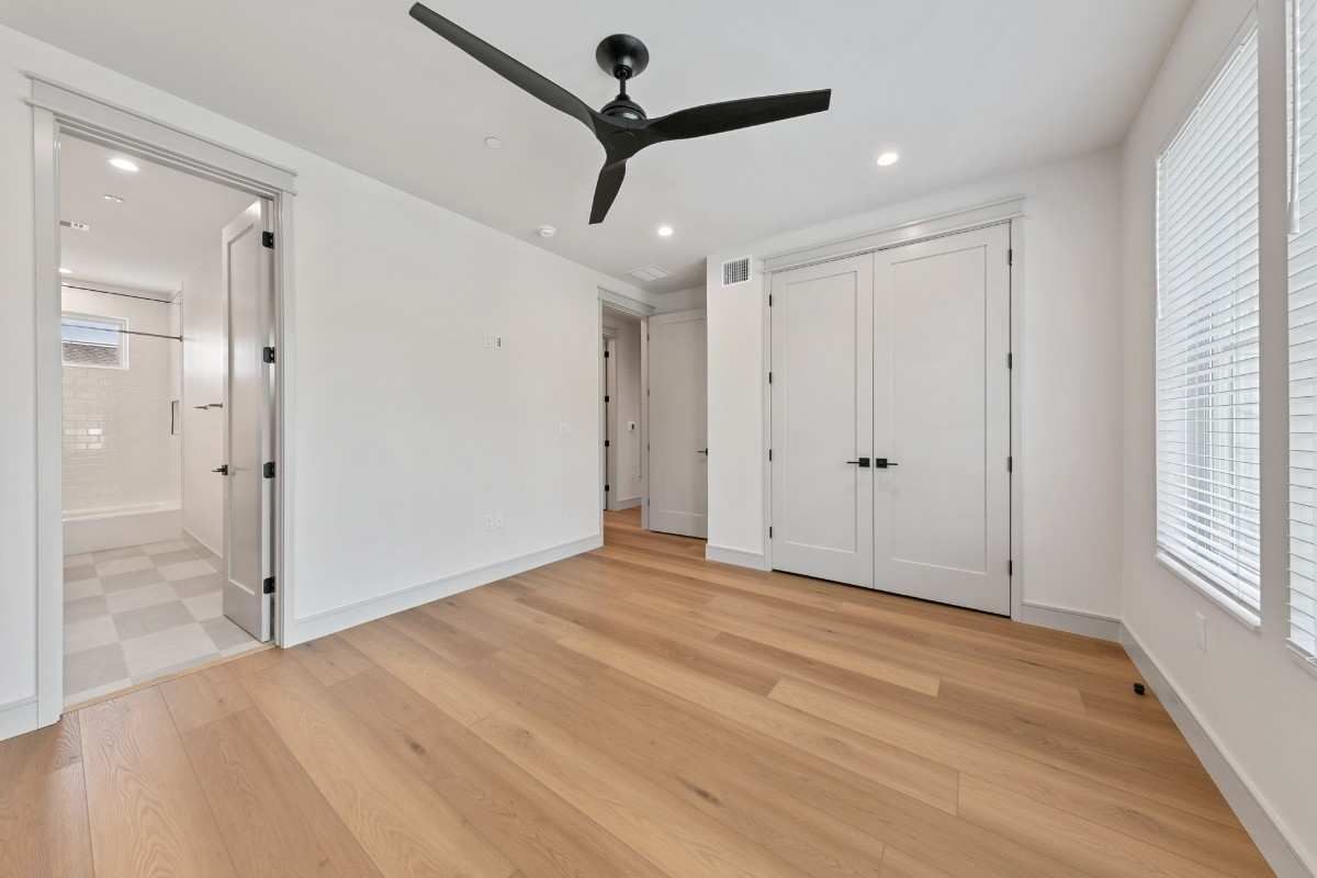 Empty room with hardwood floors, white walls, black ceiling fan, and bathroom door.