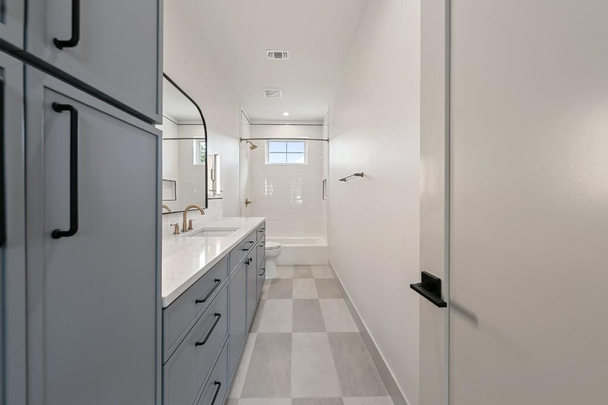 Long, narrow bathroom with gray cabinets, a white countertop, and checkered floor.