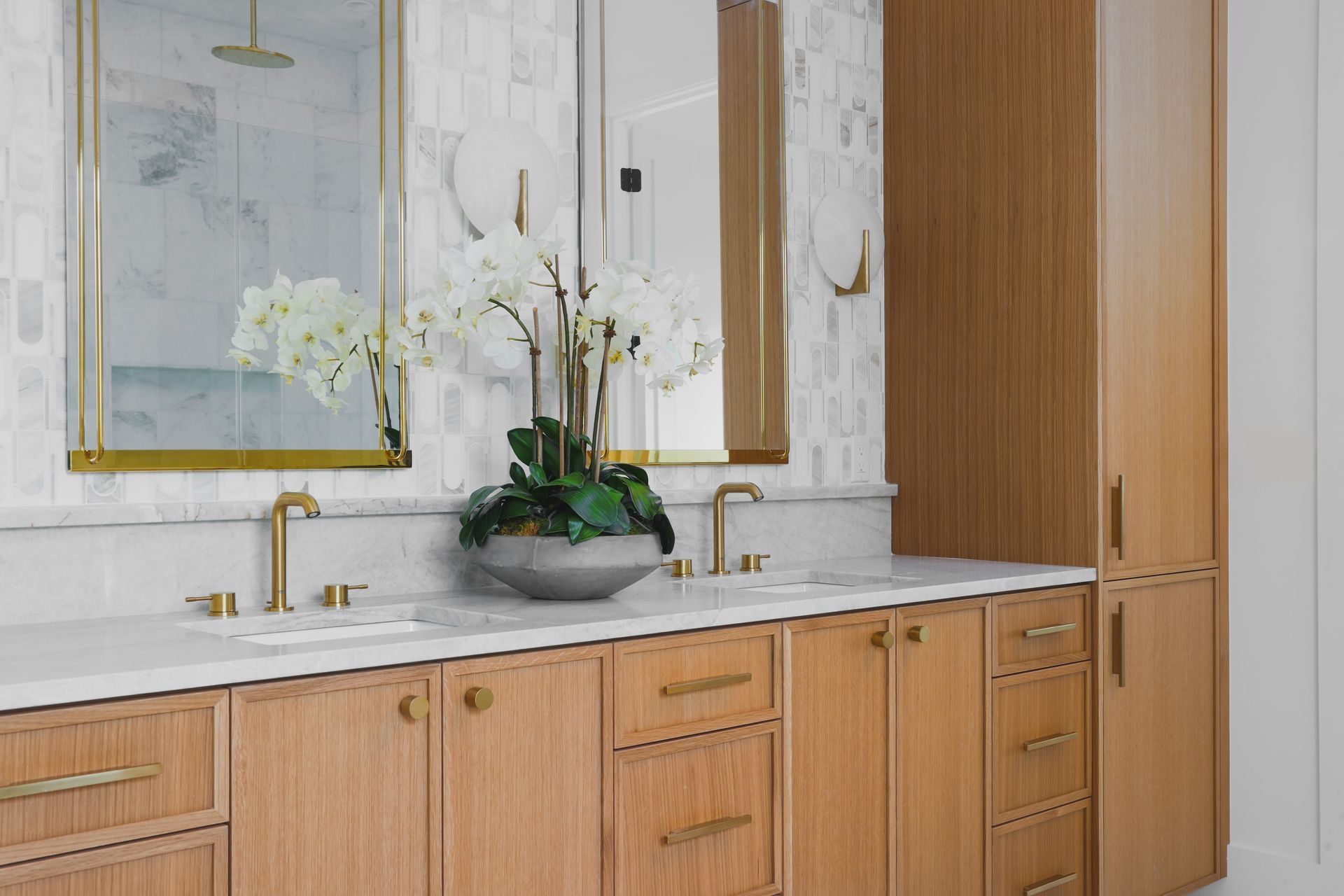 Wooden vanity with marble countertop, gold fixtures, mirrors, and decorative orchids in a modern bathroom.
