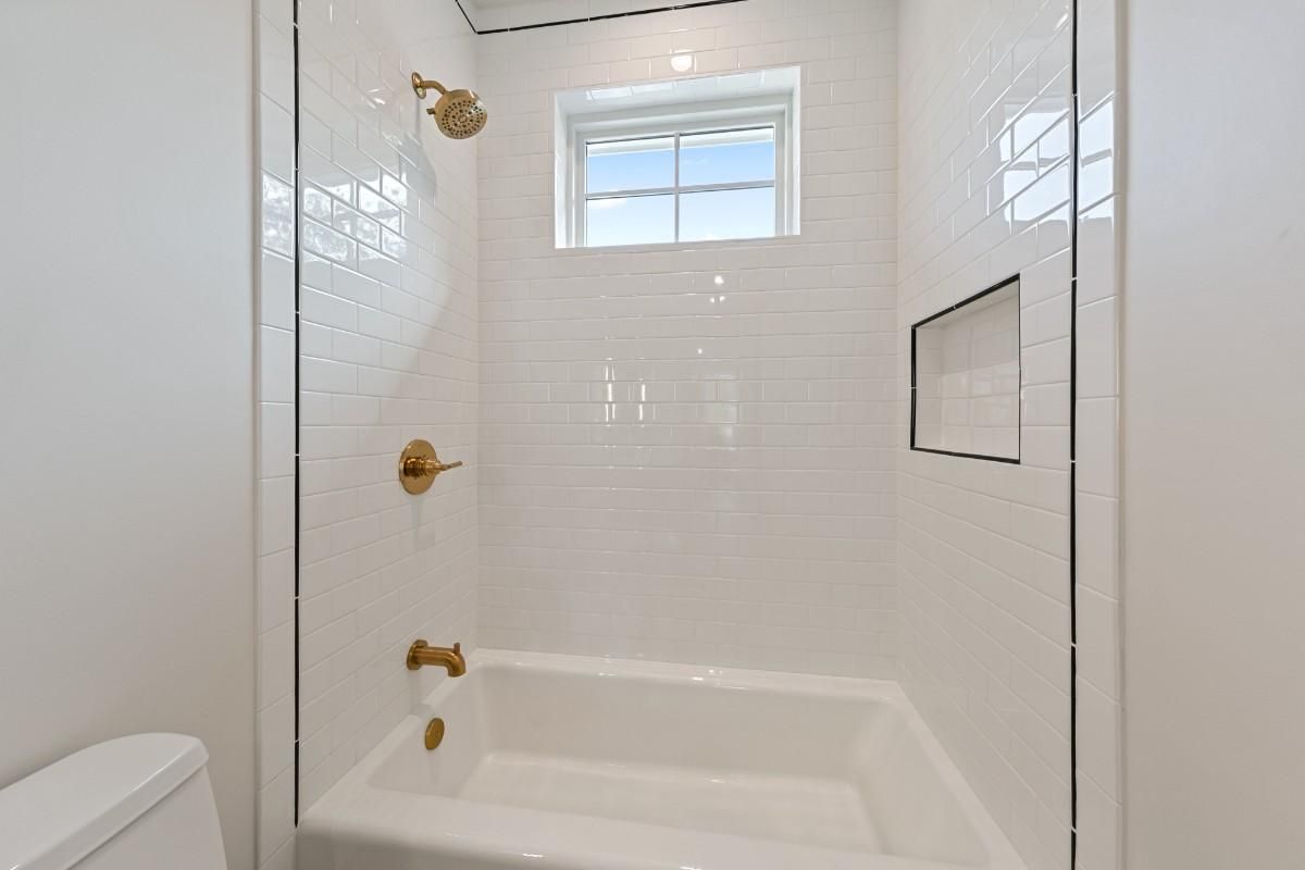 White tiled bathroom with gold fixtures, including a showerhead, faucet, and tub. A window is above the tub.