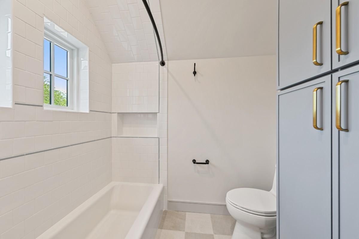 White bathroom with a tub, toilet, and cabinet with gold handles; a small window.
